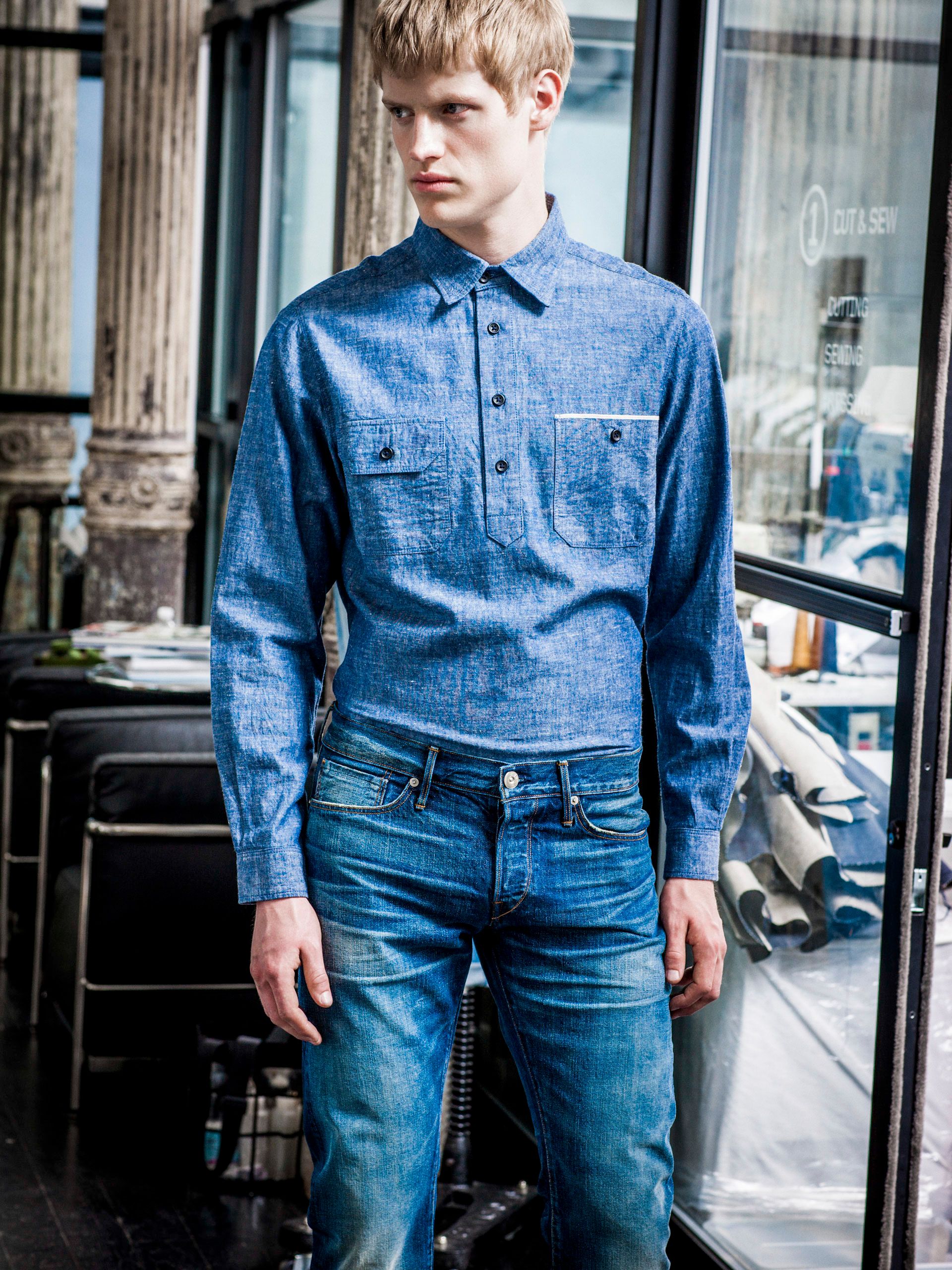 Man wearing blue chambray pullover shirt and slim fit jeans, standing outdoors in urban setting.