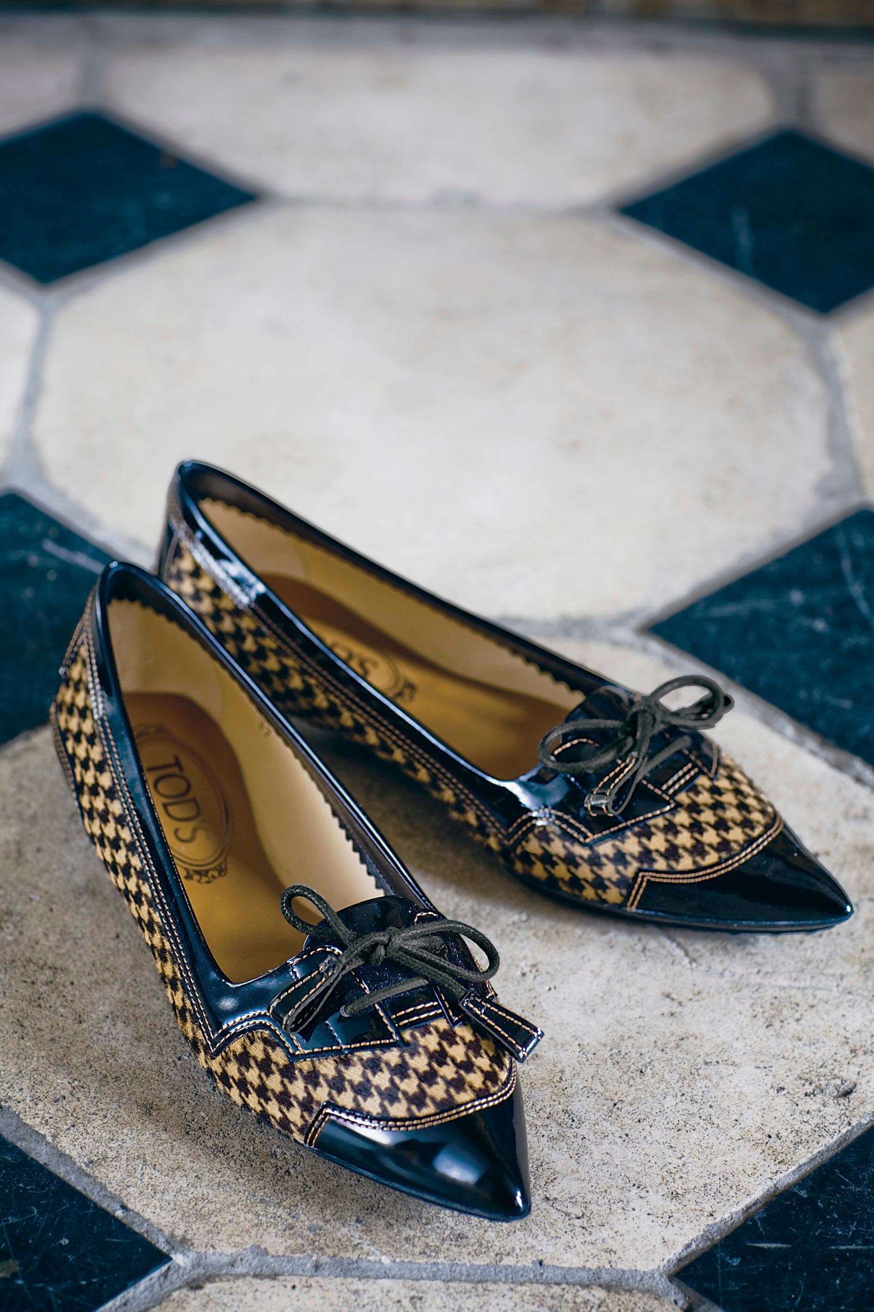 Tod's pointed-toe checkered flats with black patent leather trim and bow tassels displayed on decorative tile flooring.