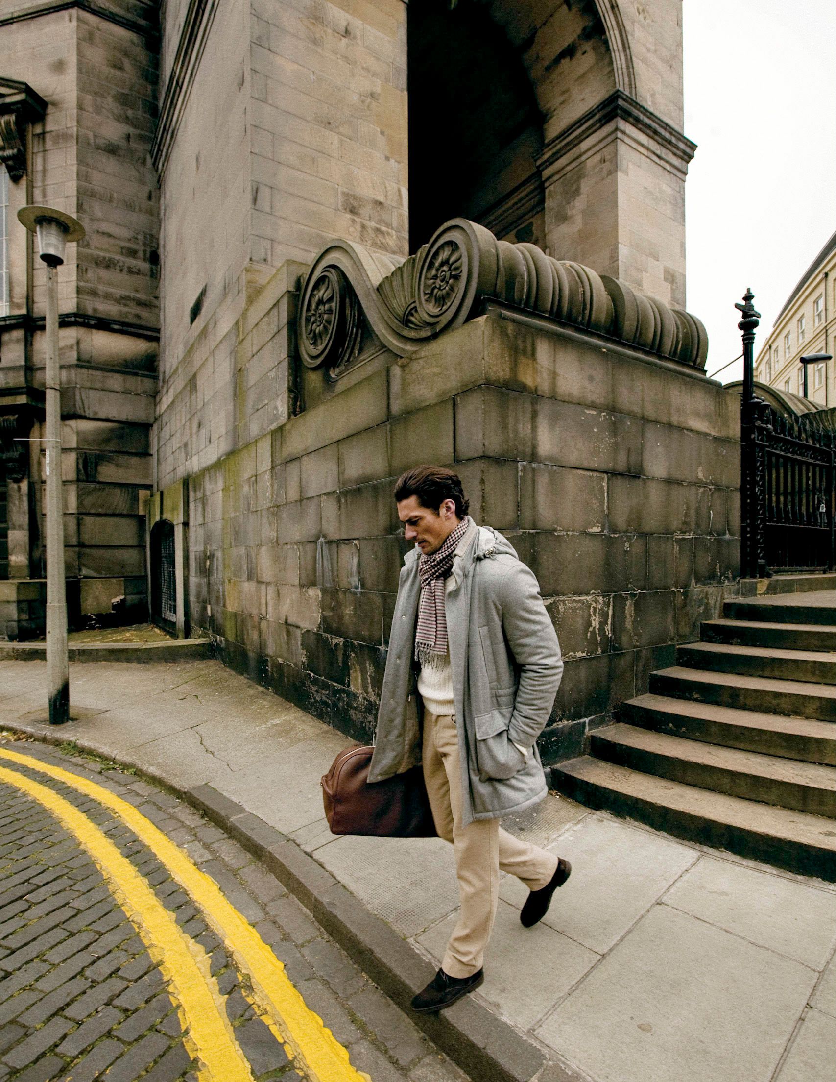 Stylish man in gray coat and scarf walks past a grand stone building with ornate columns on a city street.
