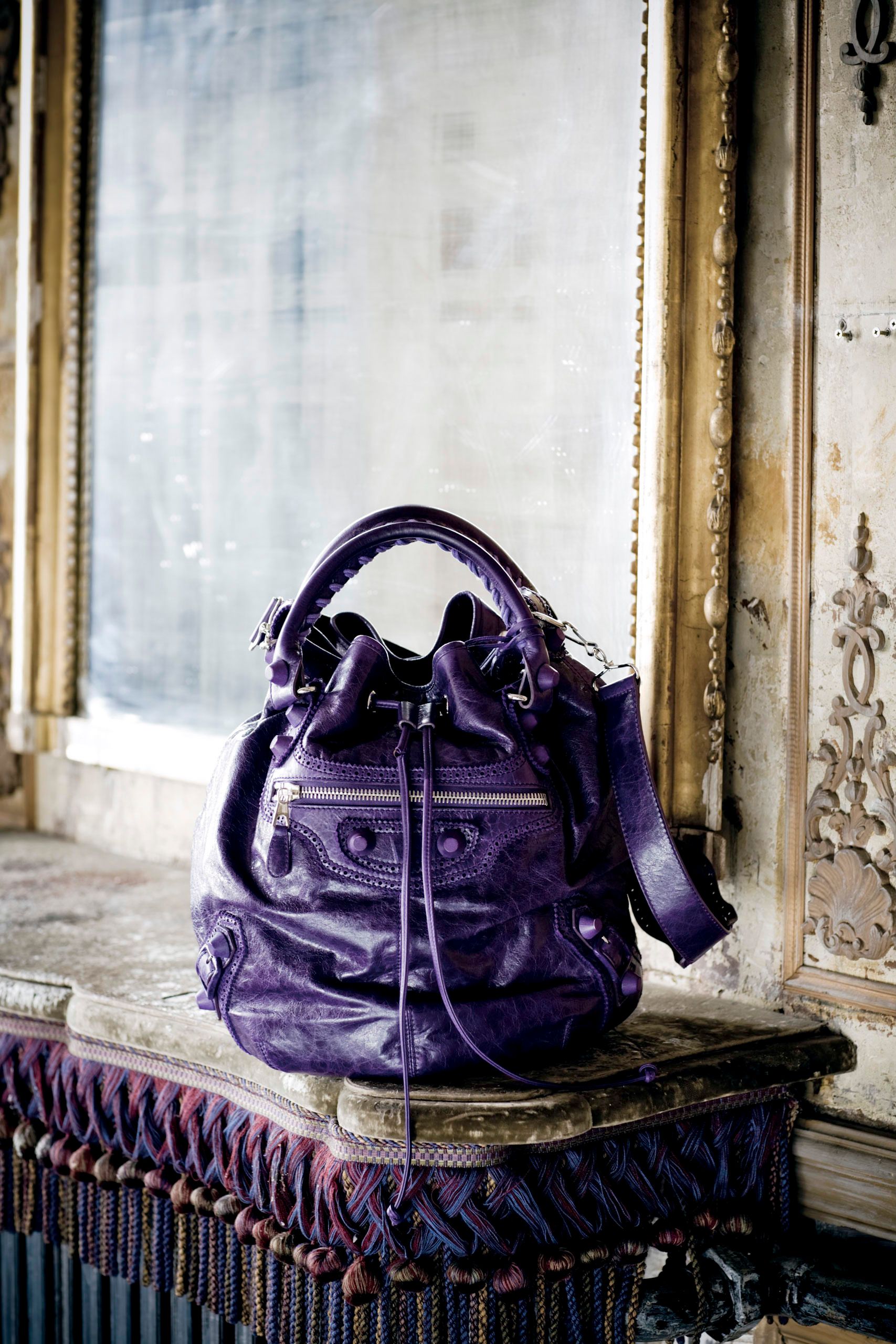 A luxurious deep purple leather handbag with drawstring detail sits on an ornate vintage surface near a gilded mirror.