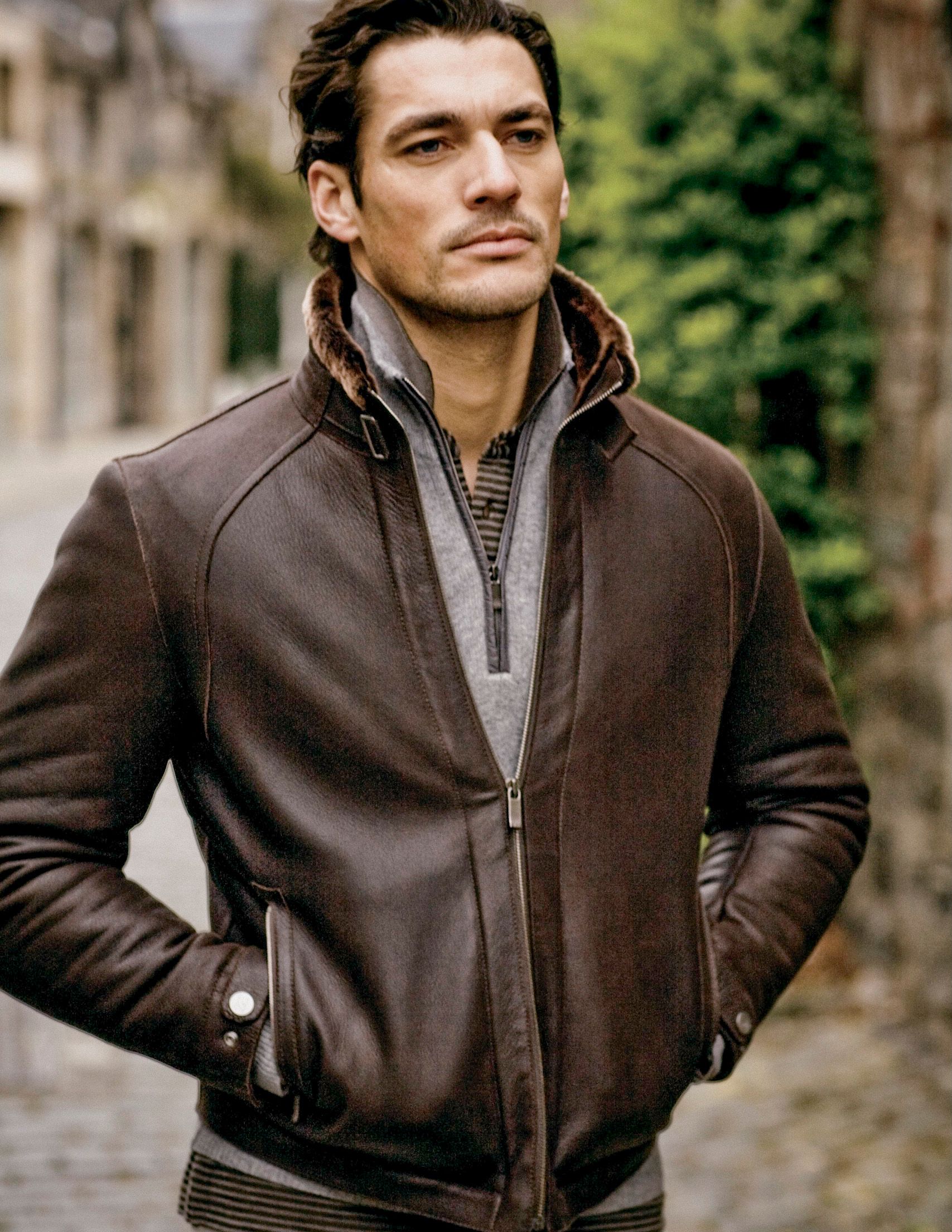 Man wearing a brown leather bomber jacket over a striped sweater, standing outdoors on a cobblestone street.