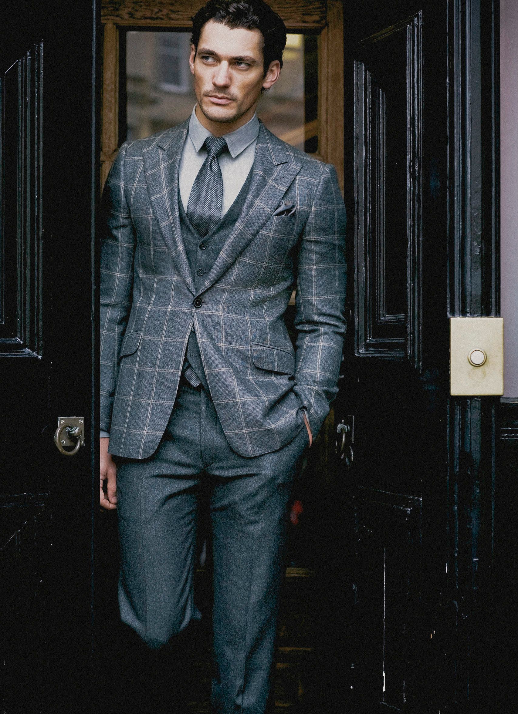 Man wearing a tailored gray windowpane check three-piece suit with tie, standing in a doorway.