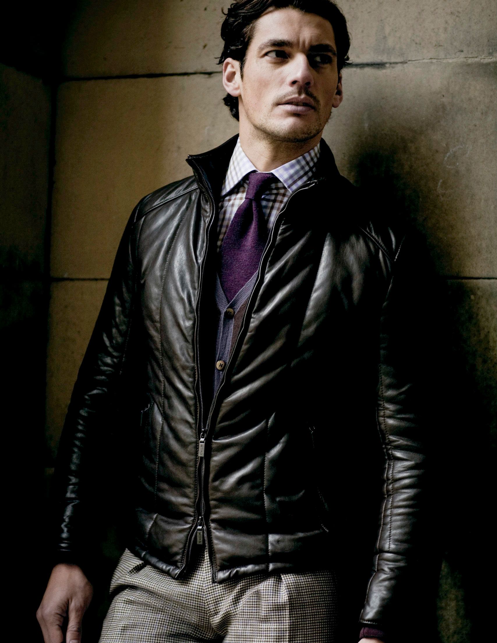 Man in black leather jacket, purple tie, and plaid shirt posing confidently in a dark, moody setting.
