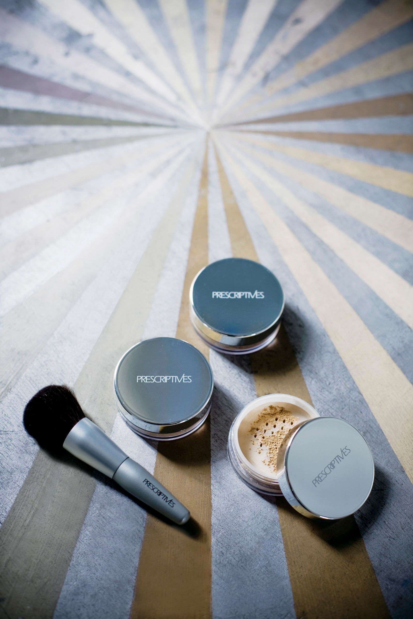 Prescriptives loose powder compacts and makeup brush displayed on striped background.