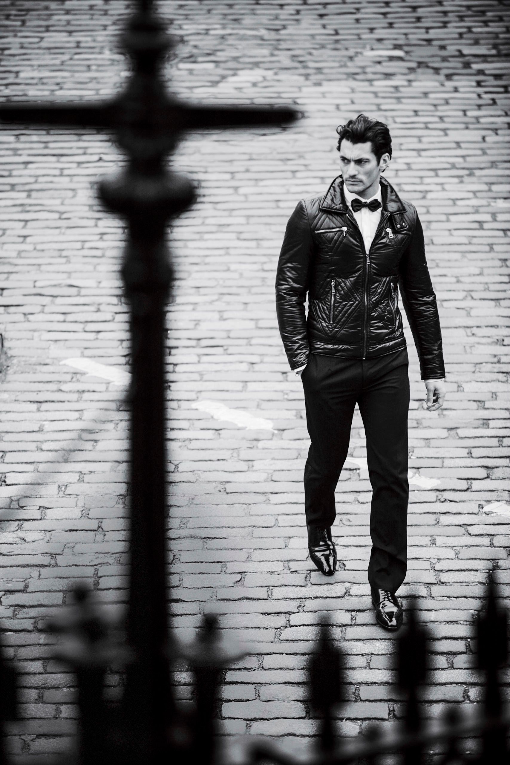 Man in black leather jacket and bow tie walking on cobblestone street in stylish monochrome fashion photo.