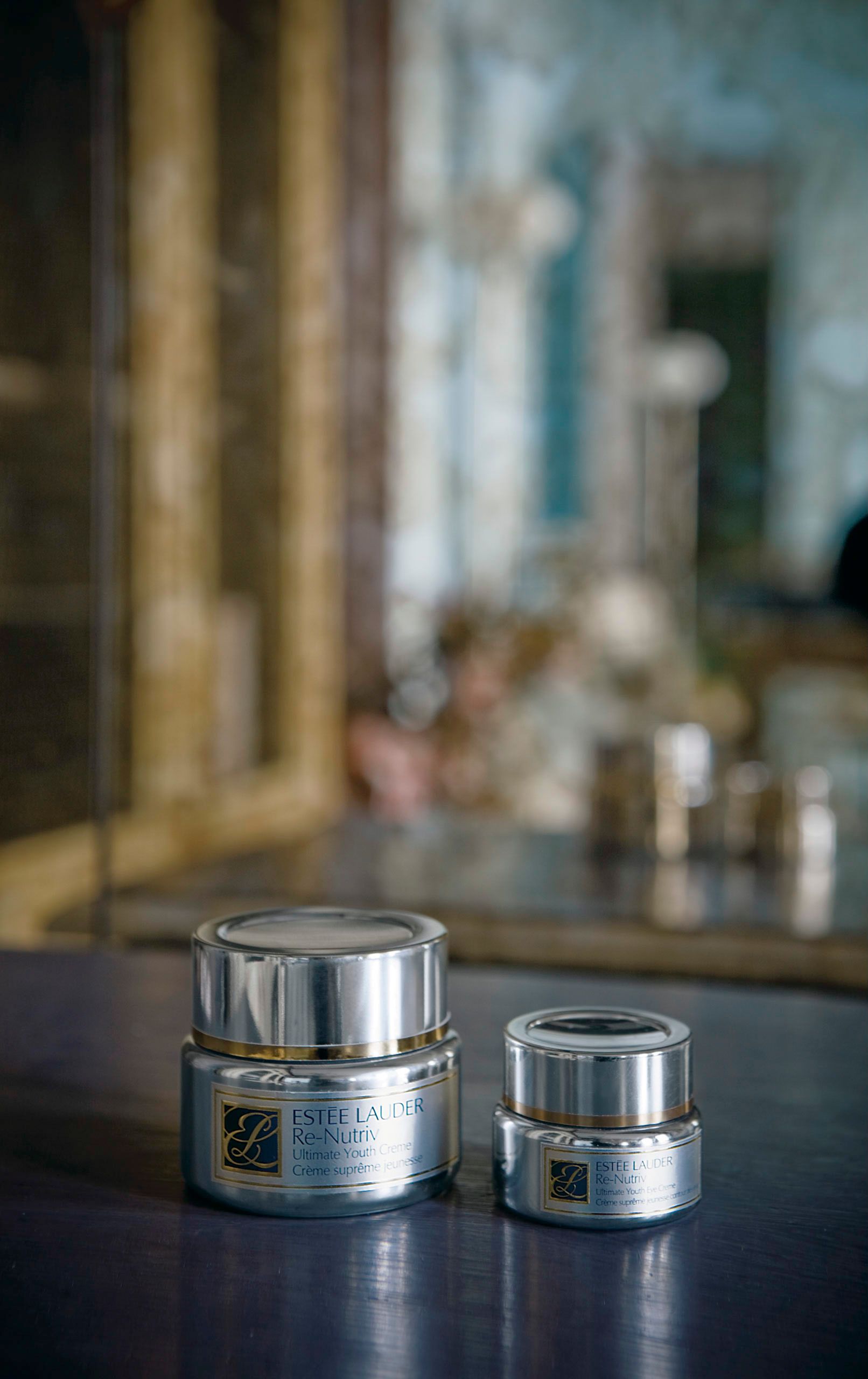 Estee Lauder Re-Nutriv skincare cream jars displayed on a reflective surface with a blurred mirror background.