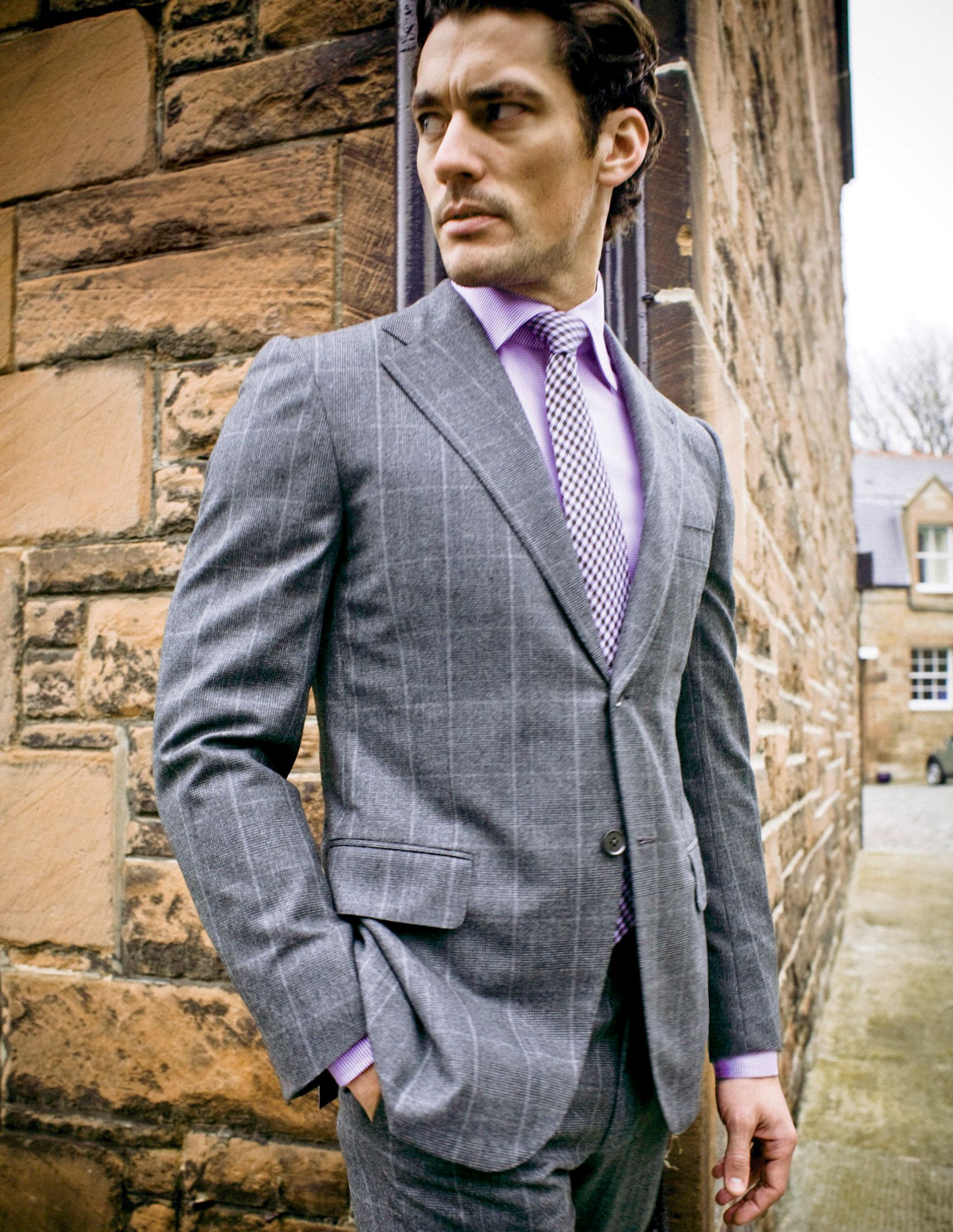 Man in gray windowpane suit with lavender shirt and patterned tie leaning against stone wall.