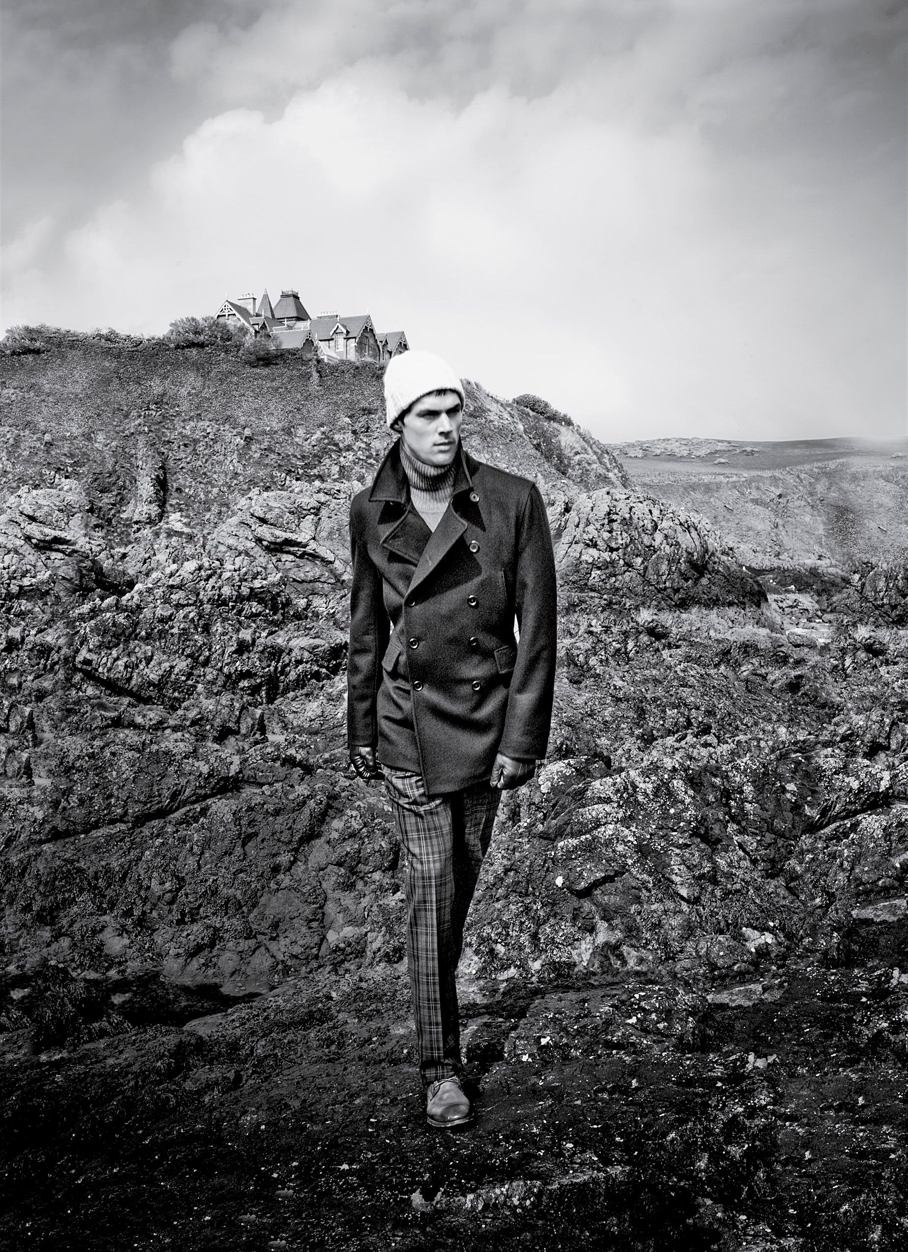 Man in peacoat and beanie stands on rocky coastal landscape in dramatic black and white fashion photography.
