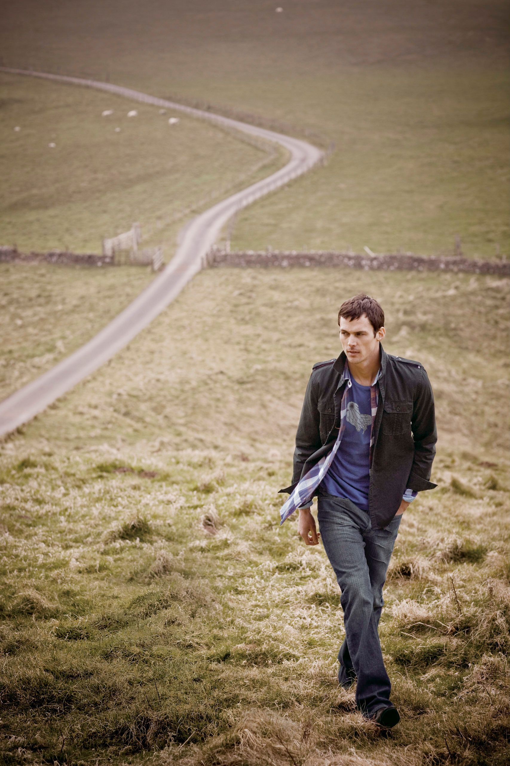 A young man in a dark jacket and jeans walks across a green grassy hillside with a winding country road behind him.