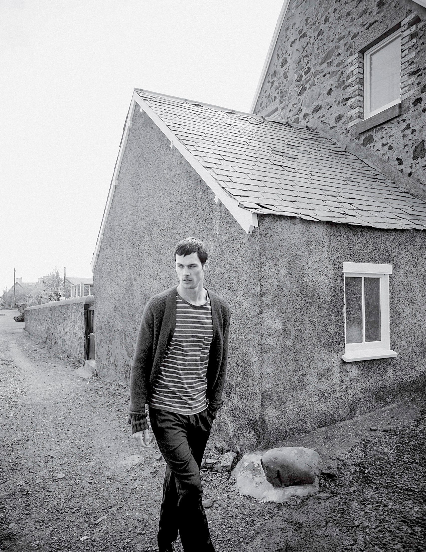 Black and white photo of a young man in striped shirt and cardigan standing on a rural path beside old stone buildings.