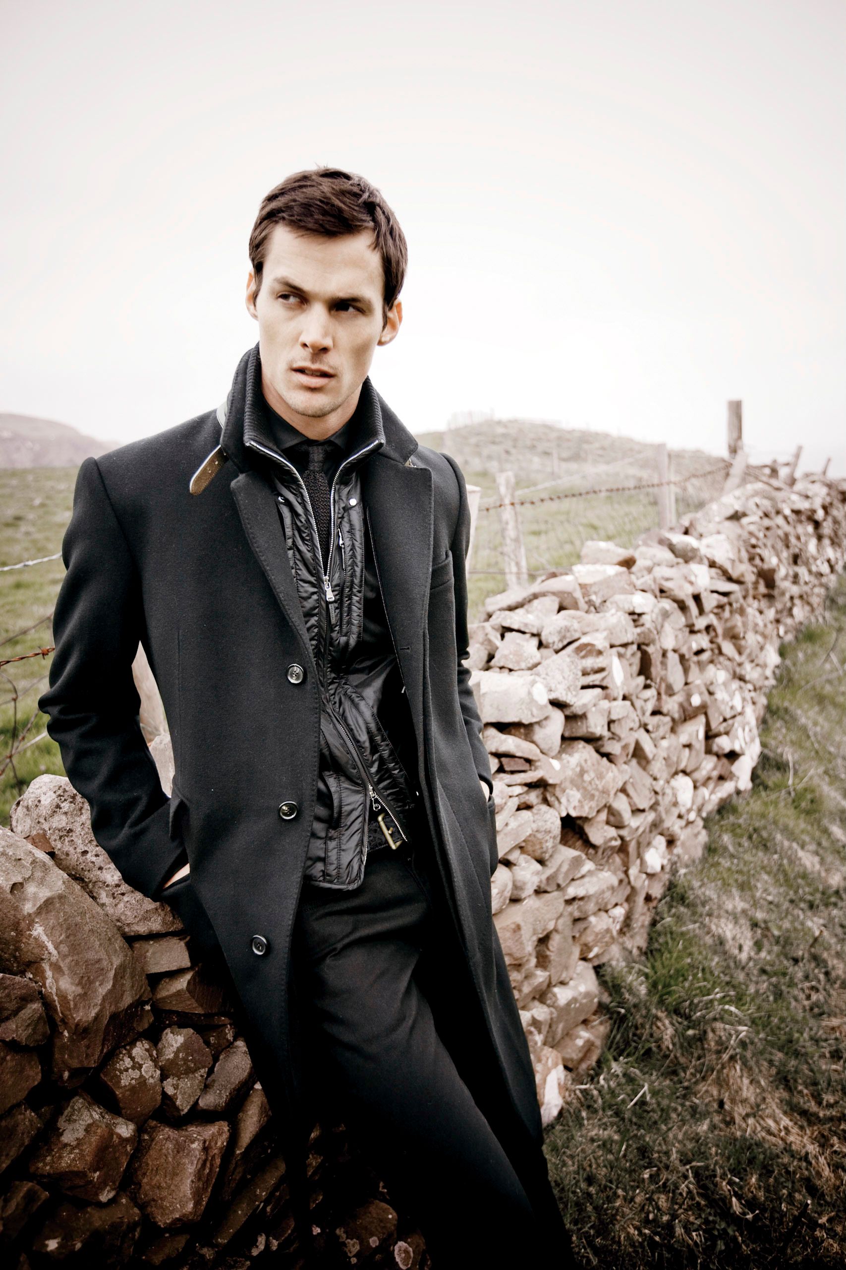 A young man in a dark long coat leans against a rustic stone wall in a moody outdoor fashion shoot.