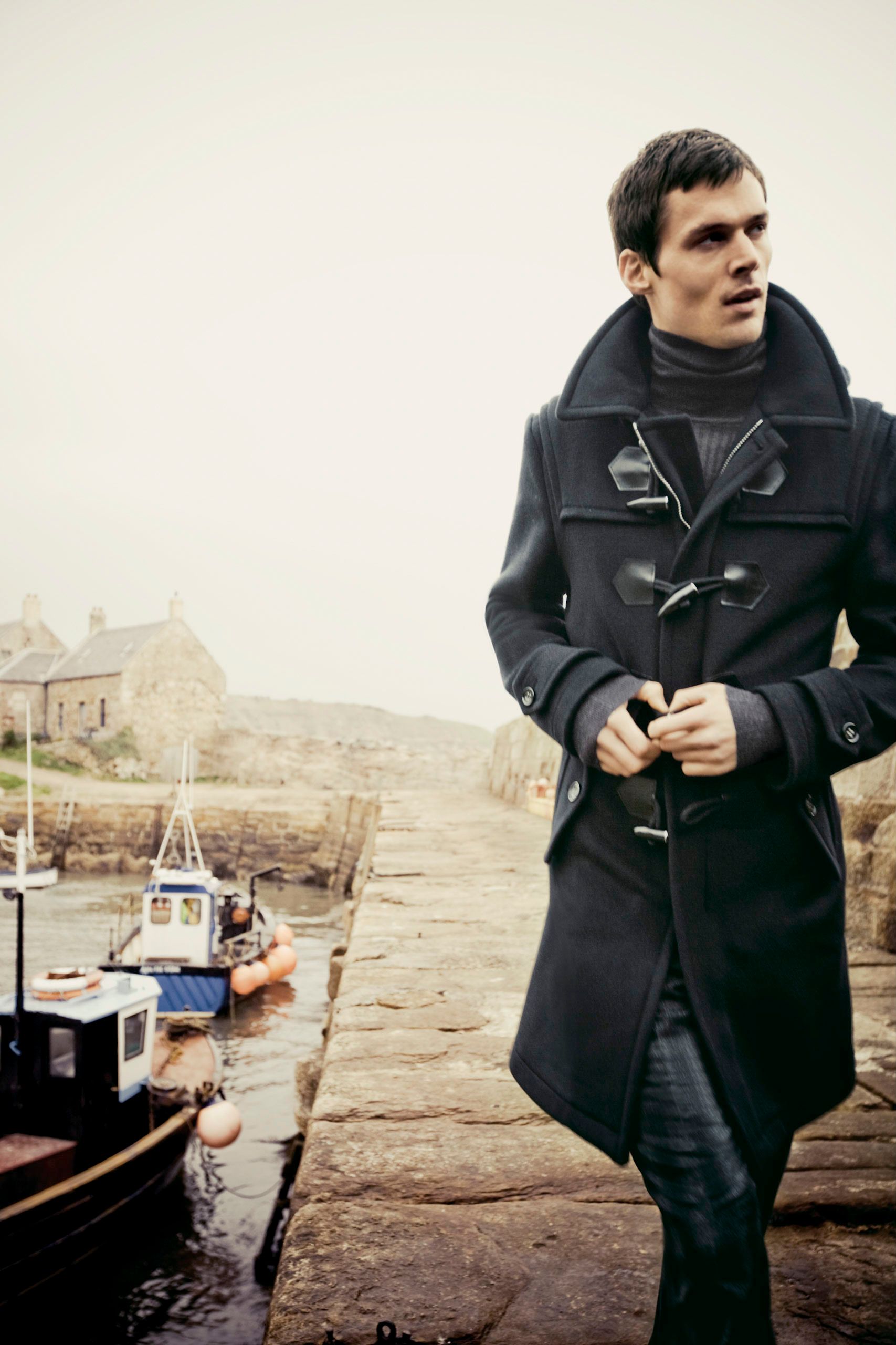 Man in dark duffle coat standing on a stone harbor pier with boats and foggy coastal village backdrop.