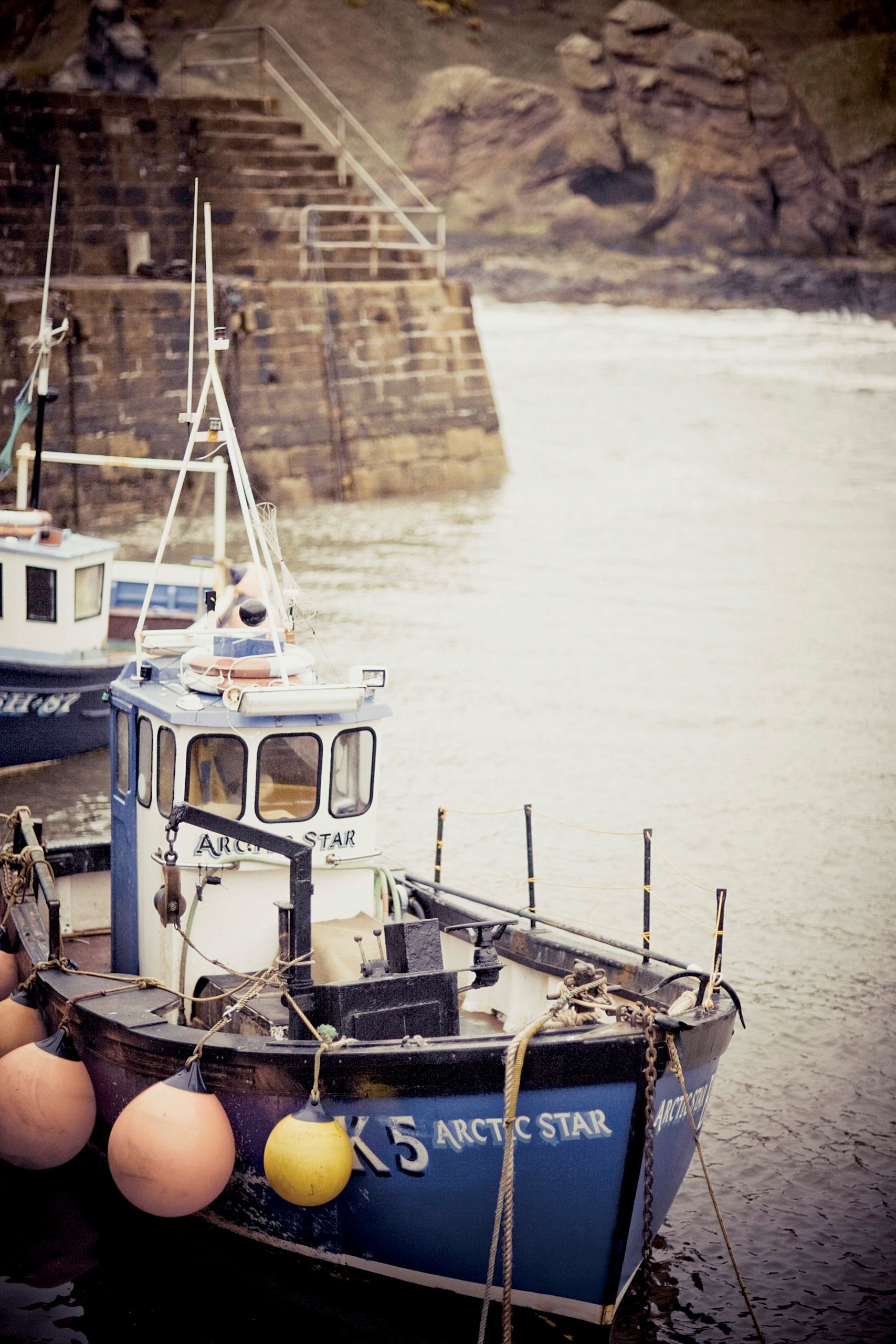 Blue fishing boat named Arctic Star K5 moored in a harbor with buoys, beside a stone wall and calm water.