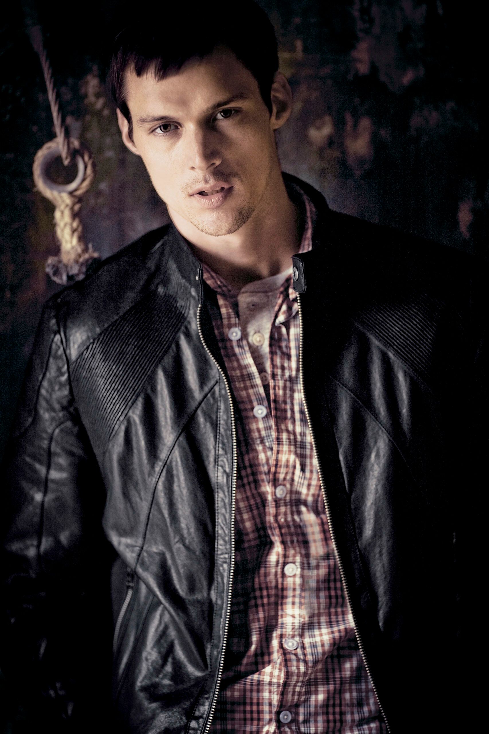 Young man in black leather jacket and plaid shirt posing against a dark textured wall.