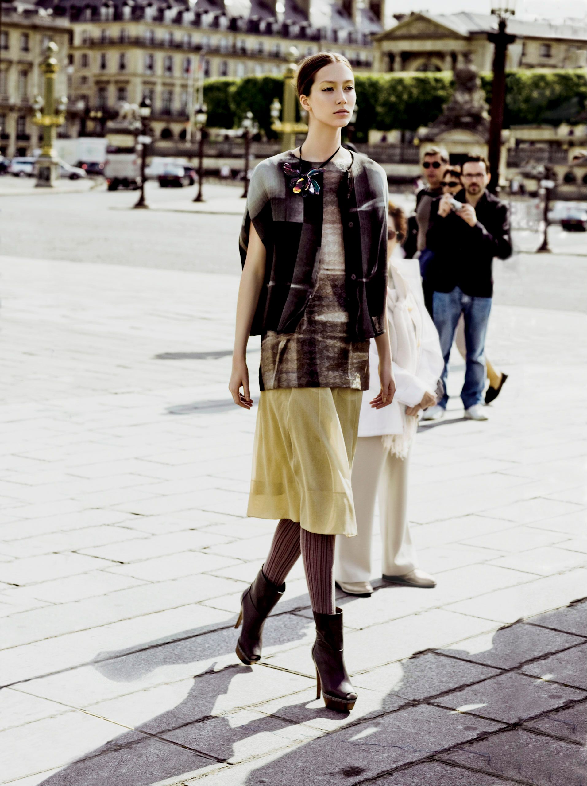 A model walks a Paris plaza in layered fashion look with plaid skirt, yellow slip dress, dark vest, and knee-high boots.