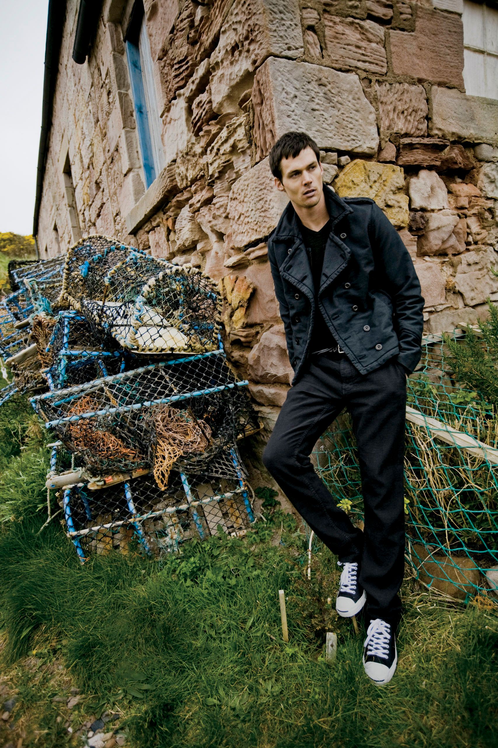Man in black jacket and trousers leaning against stone wall near lobster traps and fishing nets outdoors.