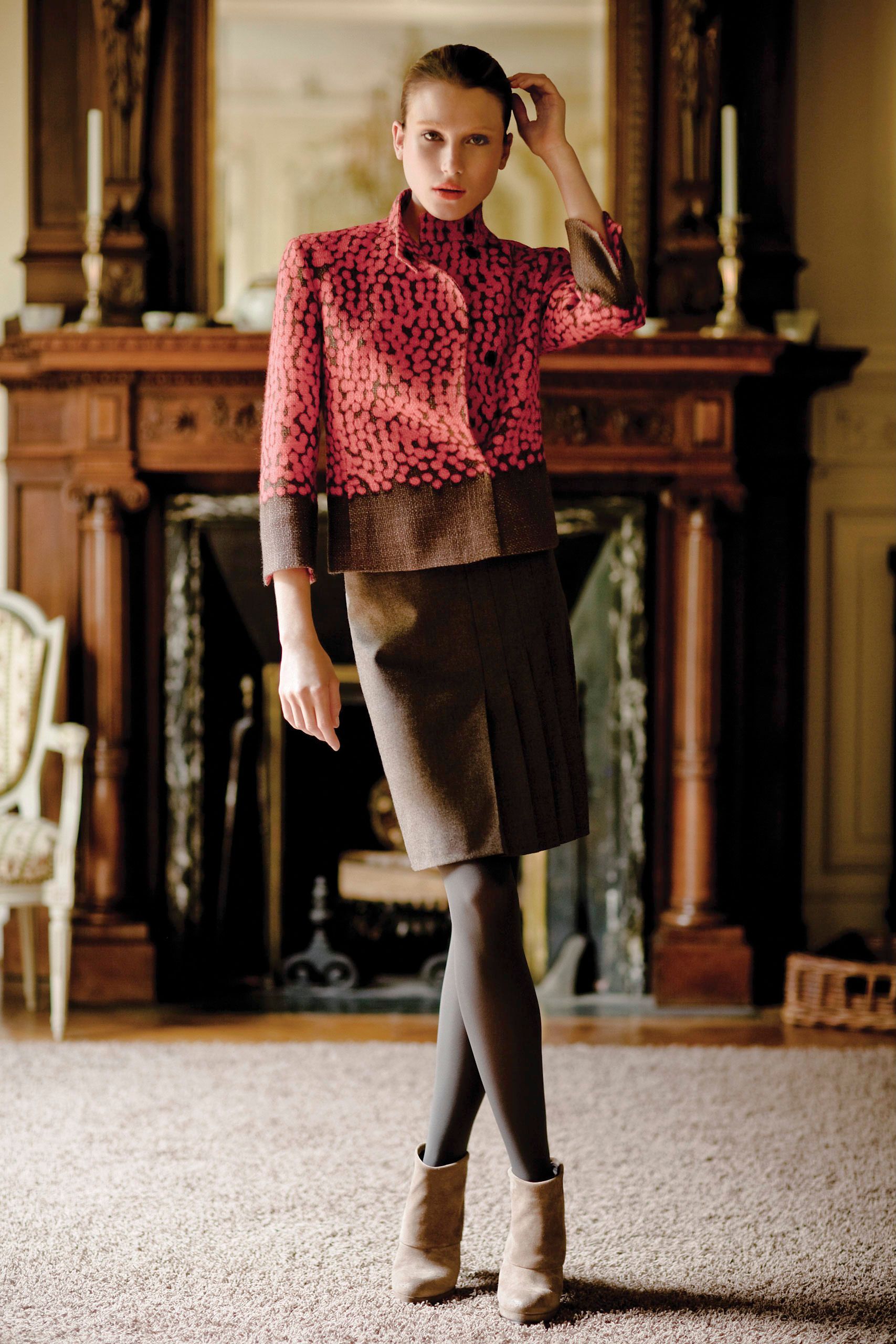 Model wearing red patterned top, brown skirt, gray tights, and beige ankle boots, posing in elegant room.