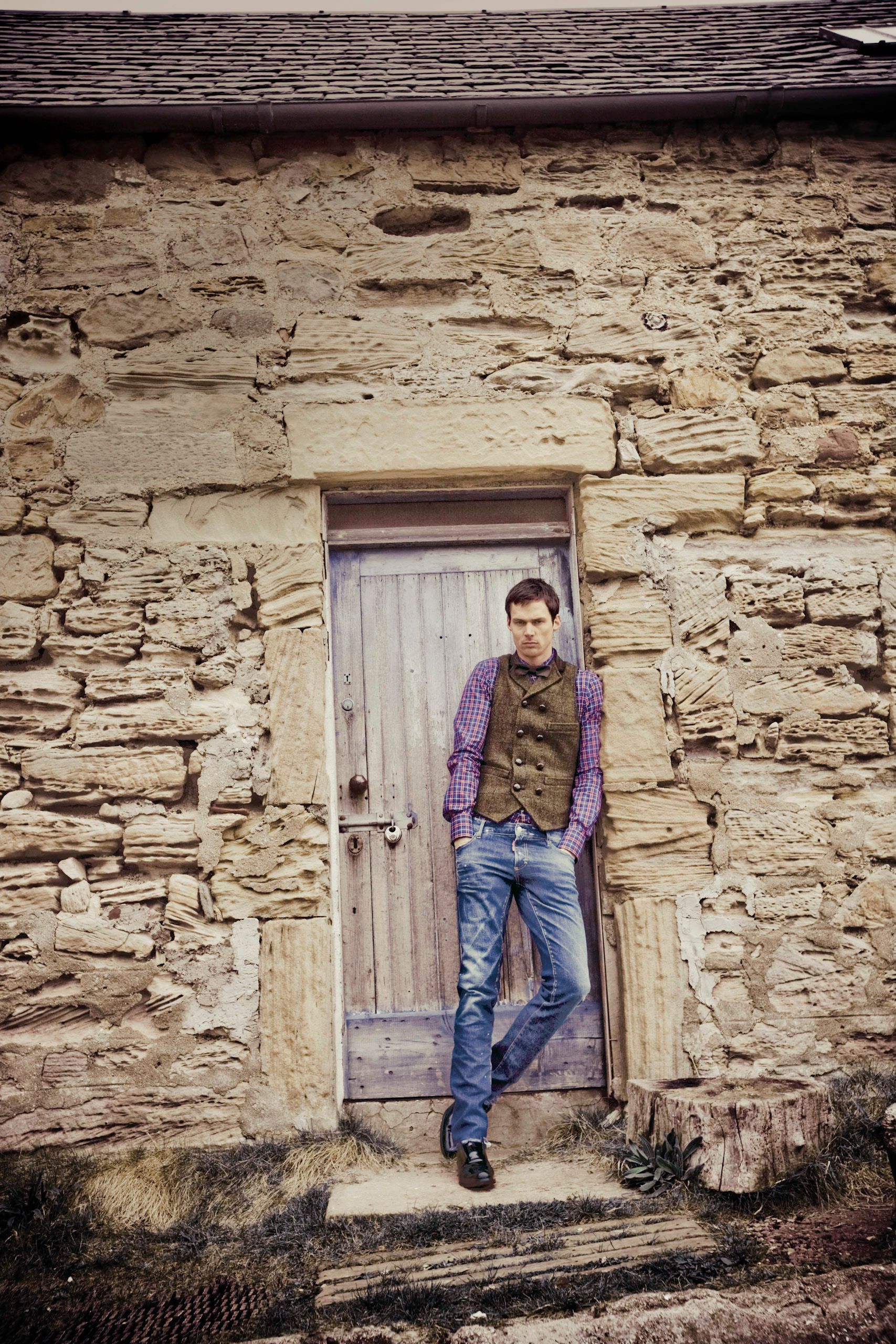 Man in vest and jeans leaning against rustic stone building wooden door.