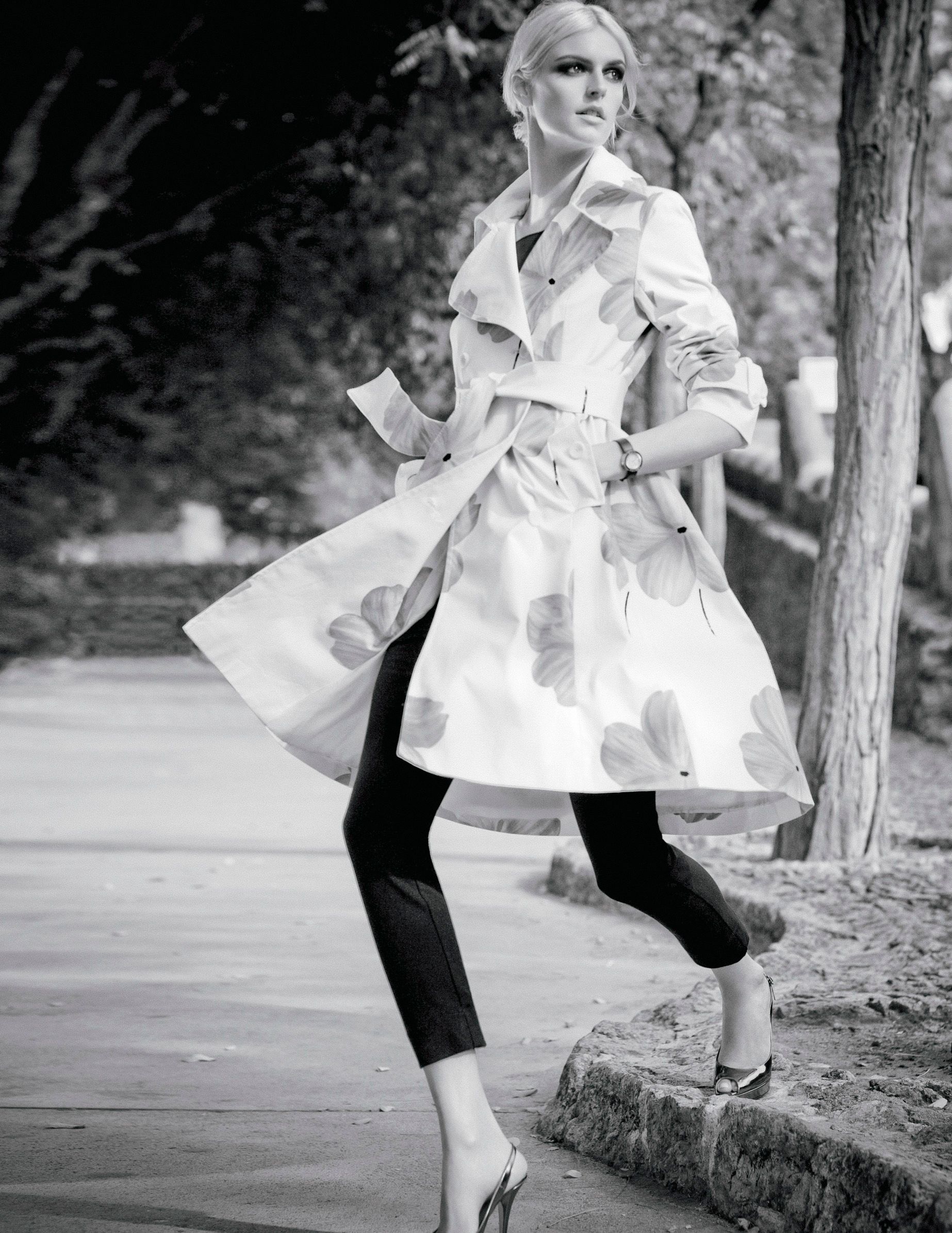 Black and white photo of a blonde woman posing in a floral trench coat with capri pants and heels outdoors.