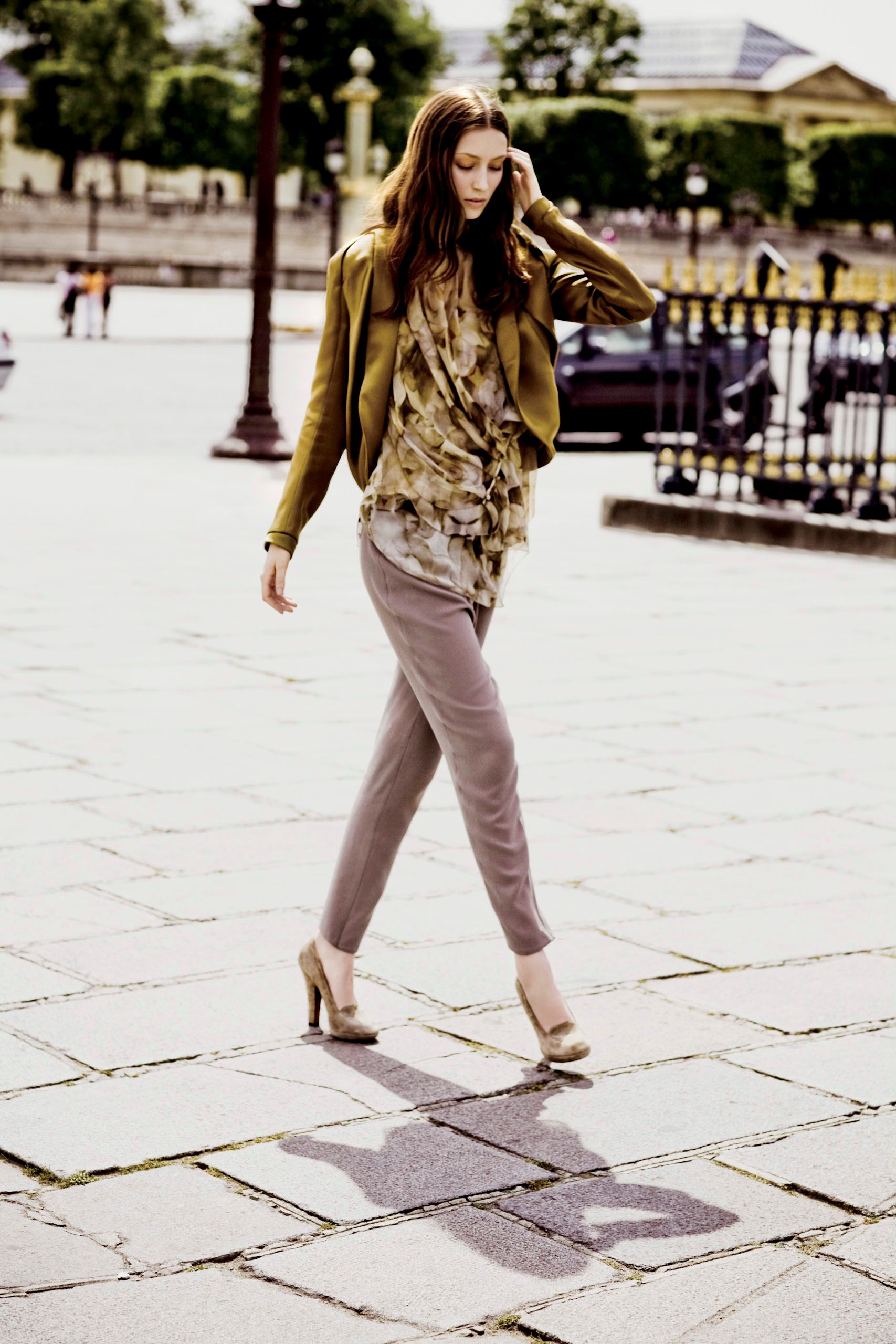 A stylish woman walks outdoors in a green jacket, floral top, taupe pants, and nude heels on a stone plaza.