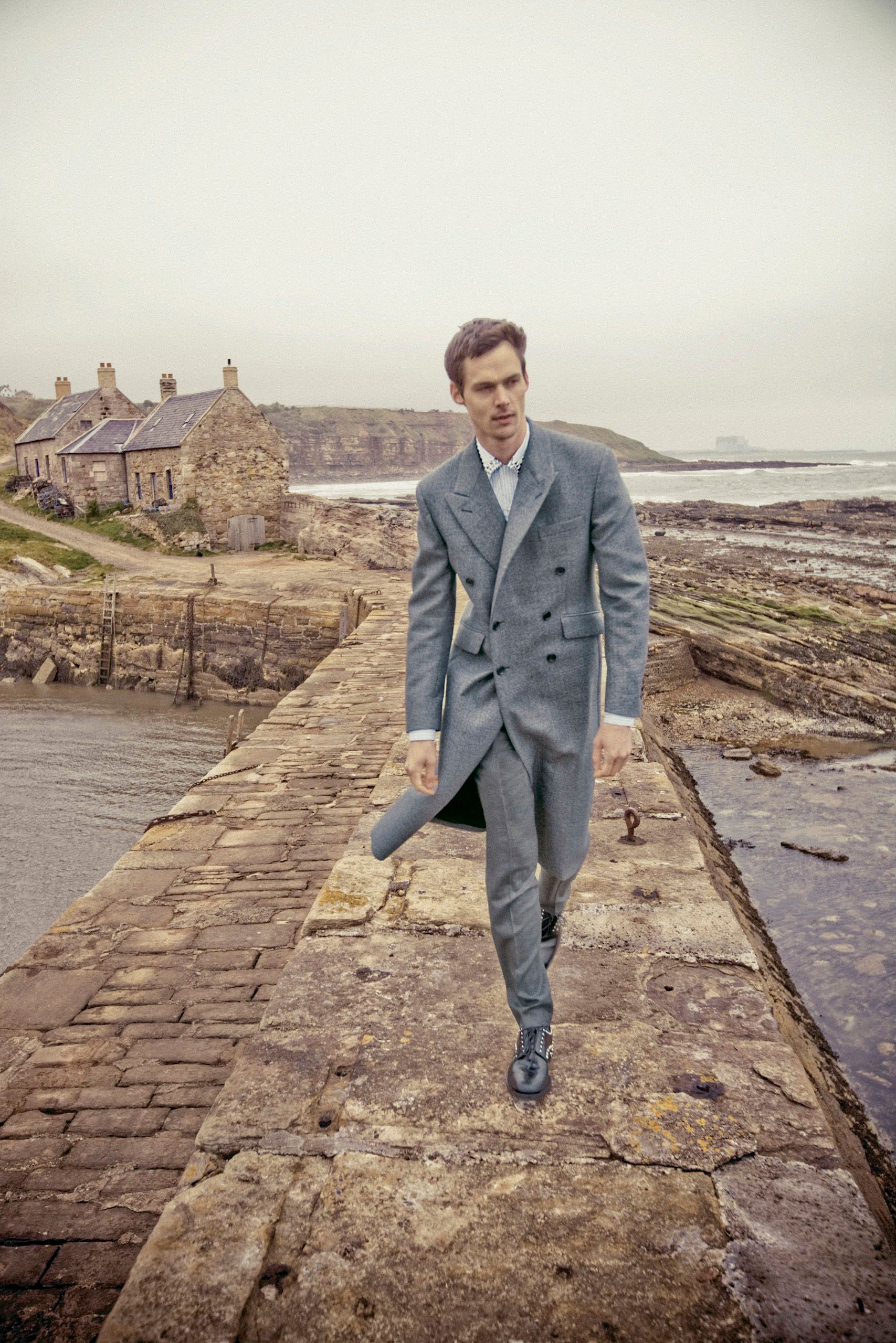 Man in grey double-breasted suit walking on stone pier with coastal village and rocky shoreline in background.