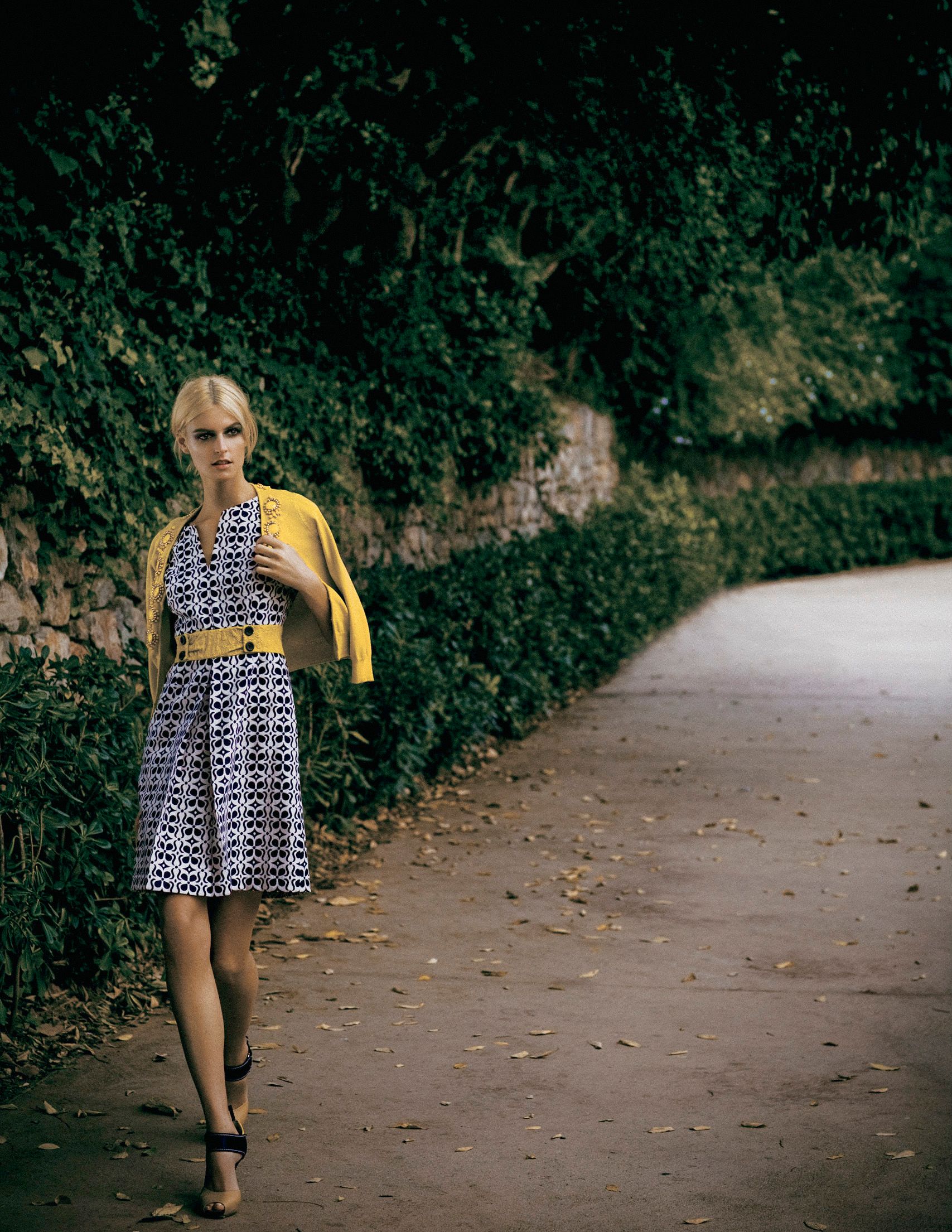 Woman in patterned dress with yellow cardigan and belt walking on a tree-lined path.