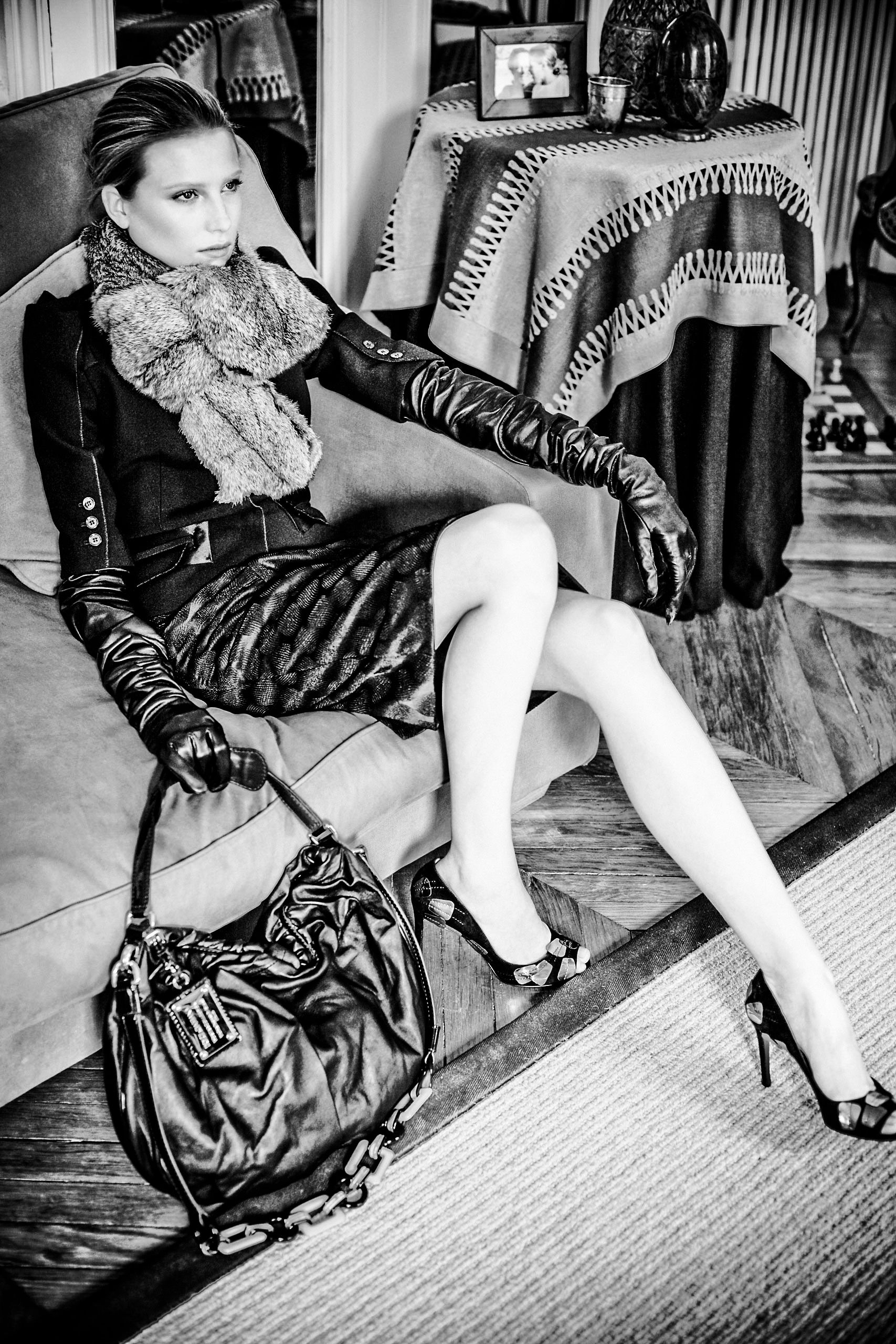 Elegant woman in black gloves, fur scarf, mini skirt, and heels poses with a leather handbag in a stylish B&W photo.