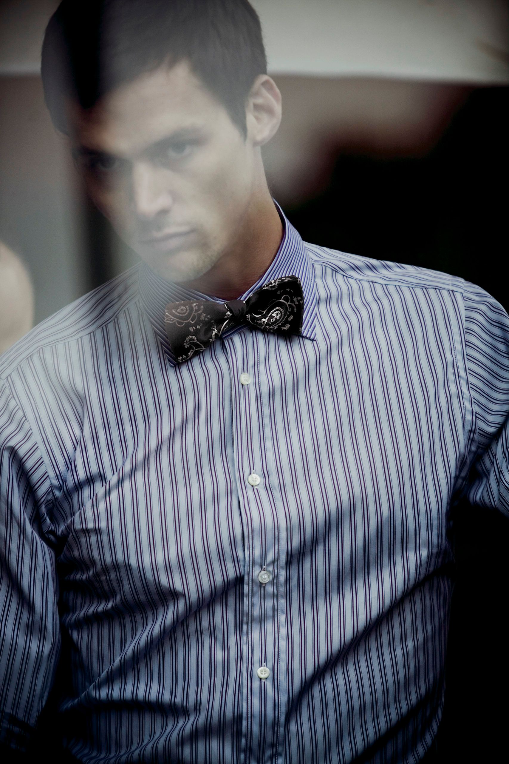 A man wearing a striped blue dress shirt with a black paisley bow tie, looking downward with a serious expression.