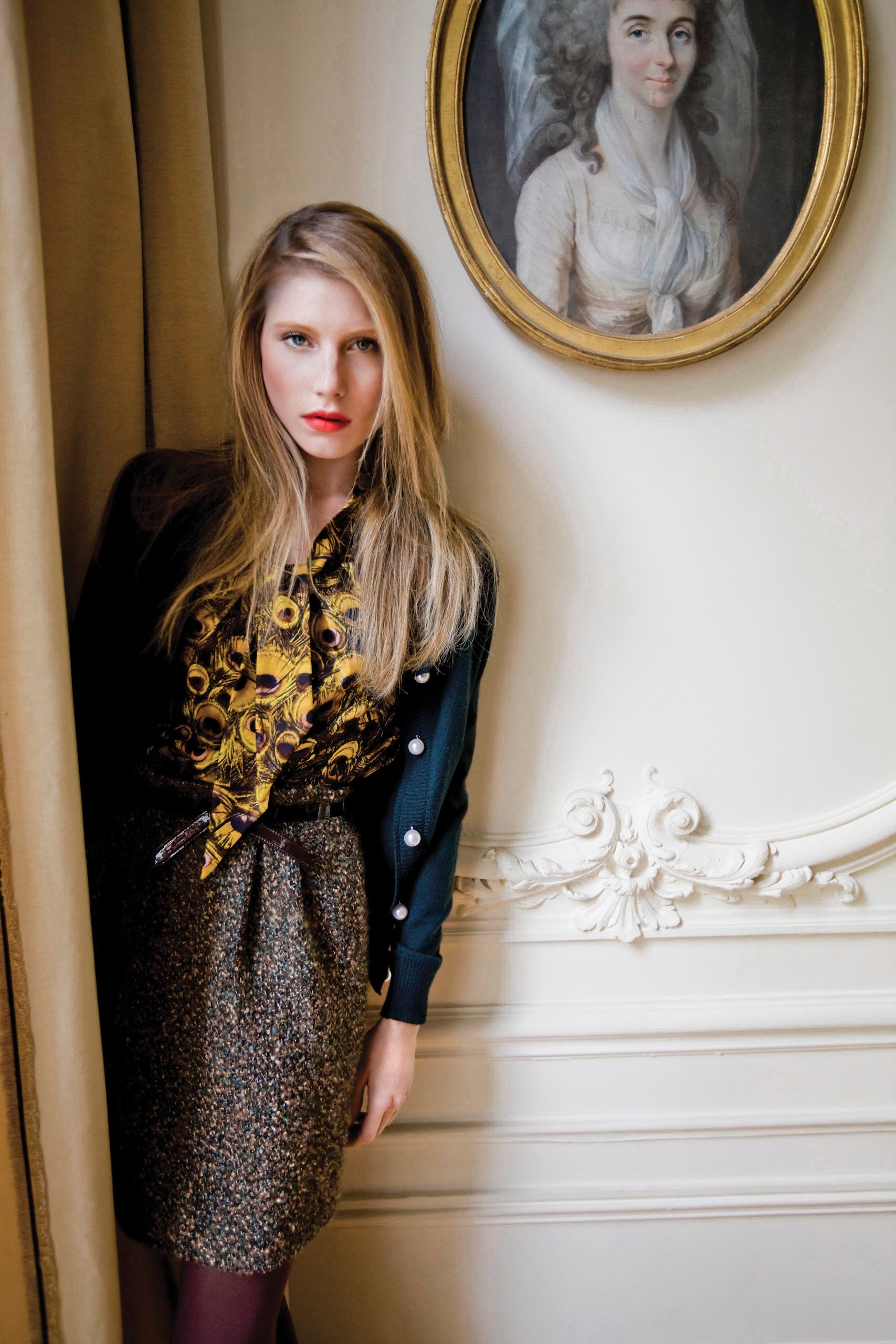 Young blonde woman in gold floral blouse and tweed skirt poses beside ornate wall molding under vintage portrait.