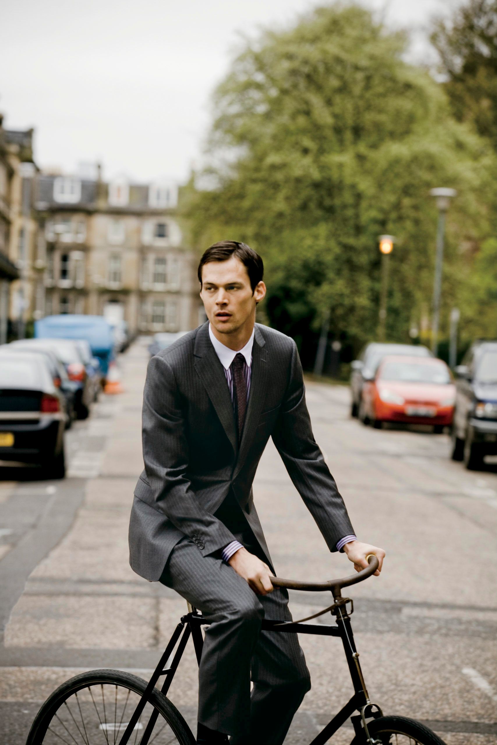 A well-dressed man in a gray suit and tie rides a black bicycle down a tree-lined urban street.
