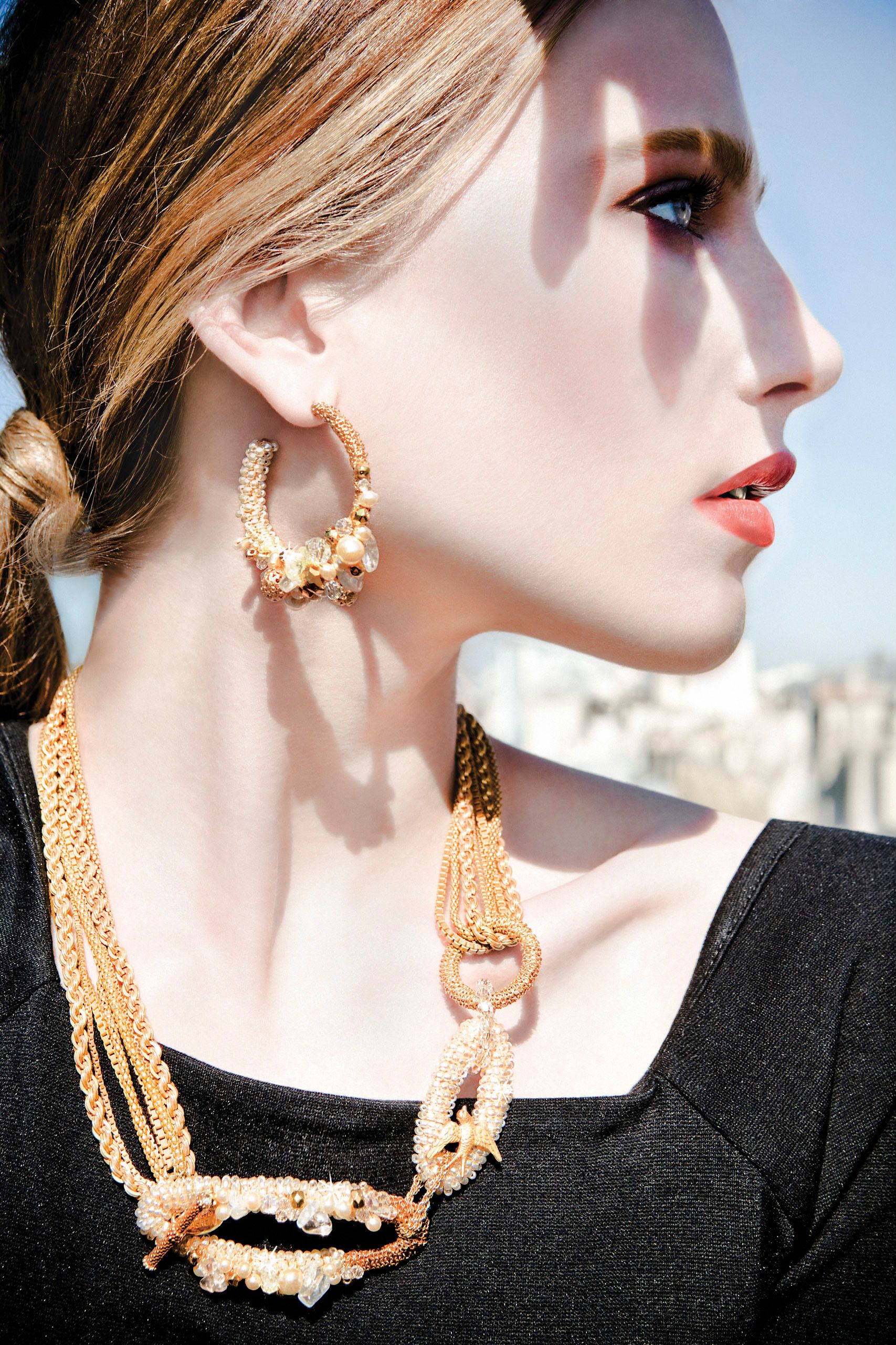 Woman in profile wearing gold hoop earrings with pearls and layered gold chain necklace against bright sky.