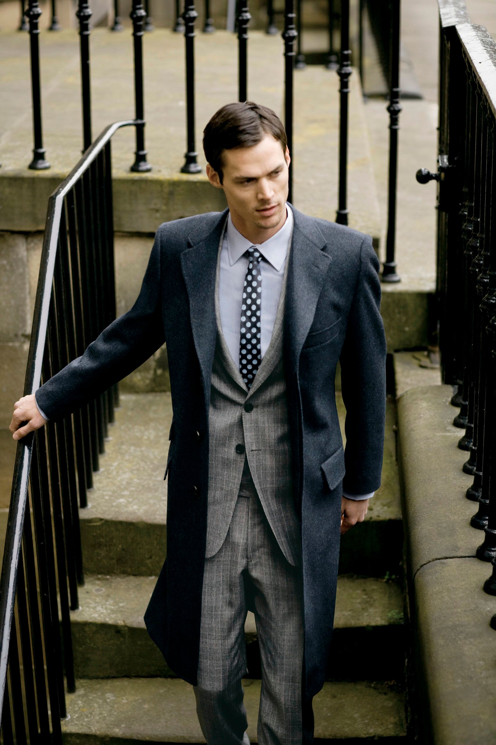 Stylish man in gray plaid suit and long coat descends stone steps holding black iron railing.