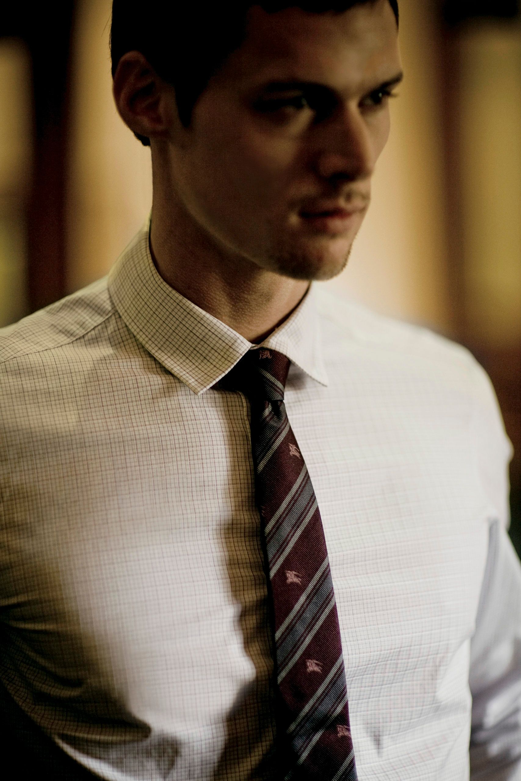 Young professional man in a checkered dress shirt and striped burgundy tie, looking away in a dimly lit setting.