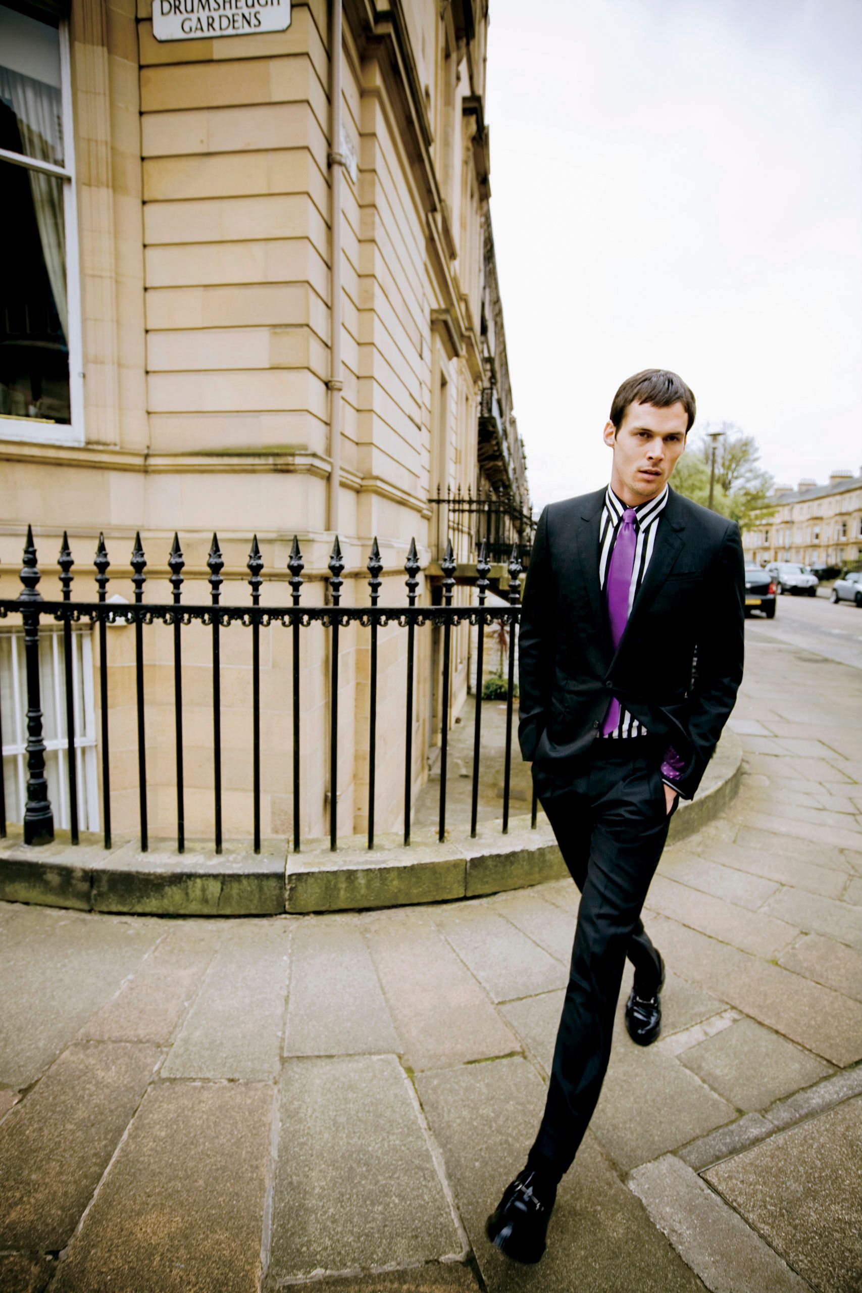 Man in black suit with purple tie walking on a city sidewalk near iron railings and a stone building.