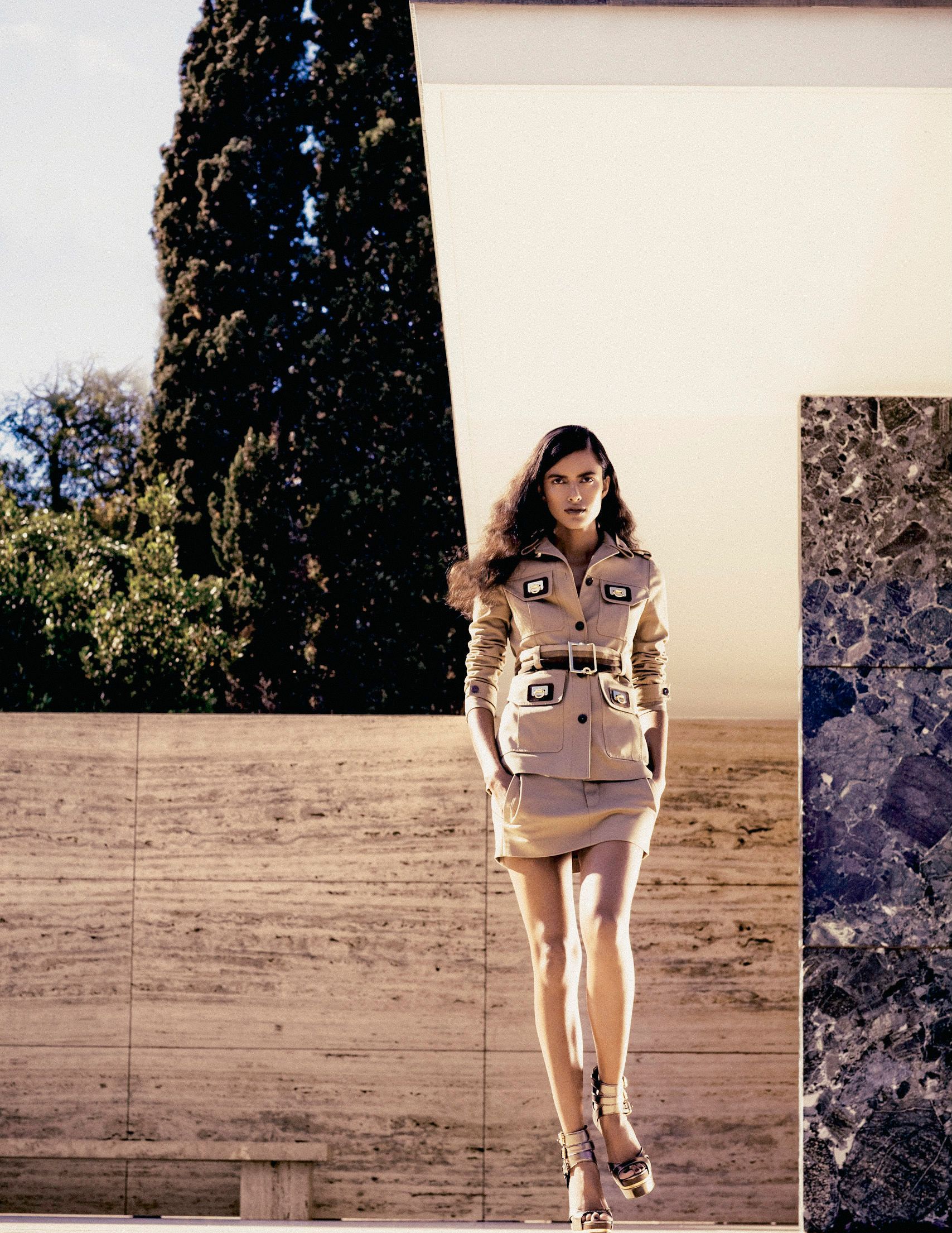 A model poses in a beige belted safari suit and heeled sandals between stone walls with a cypress tree backdrop.