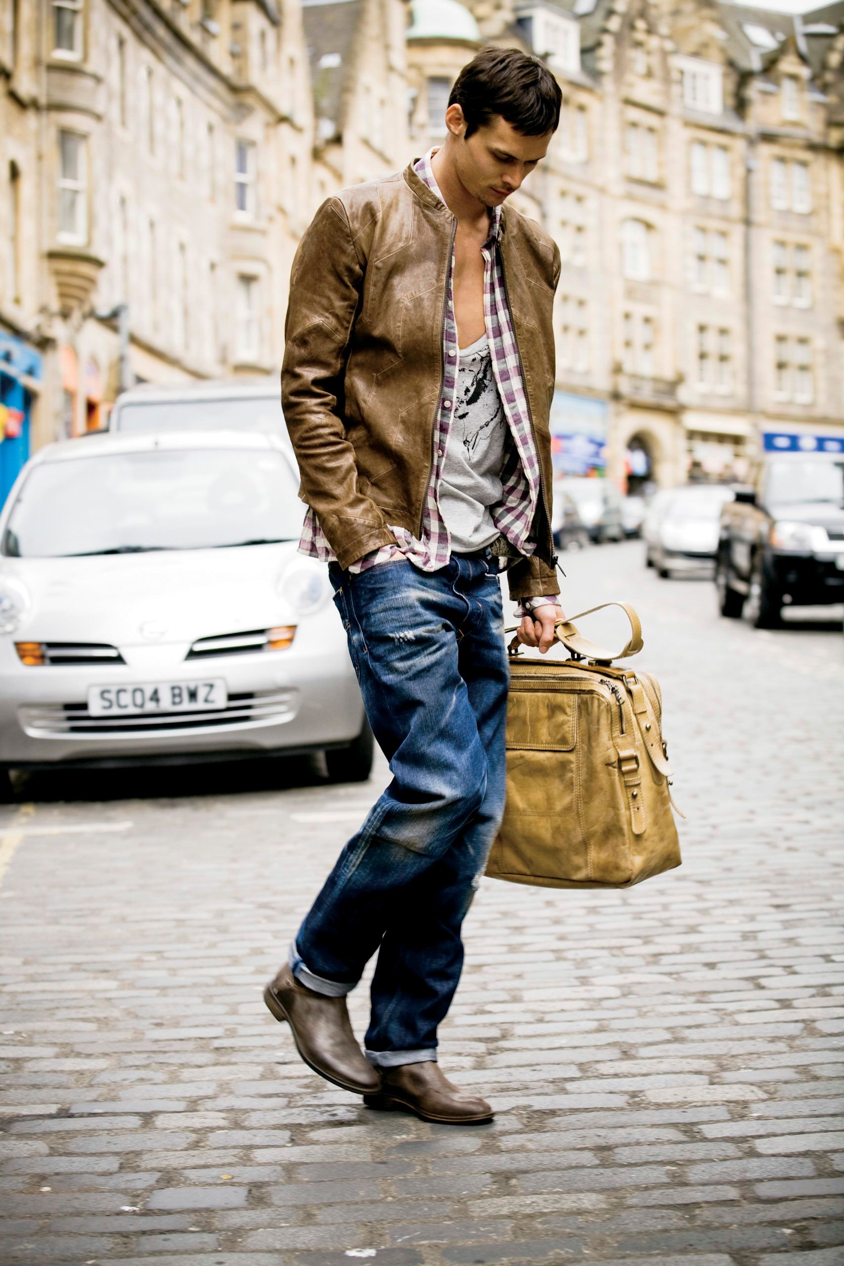 A man in jeans and brown leather jacket carries a bag on a cobblestone street.