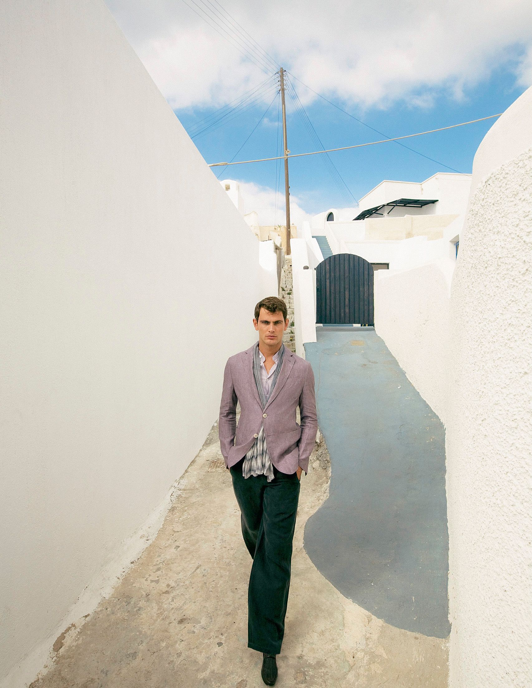 A man in a lavender blazer walks through a narrow white-walled alley under a blue sky in Santorini, Greece.