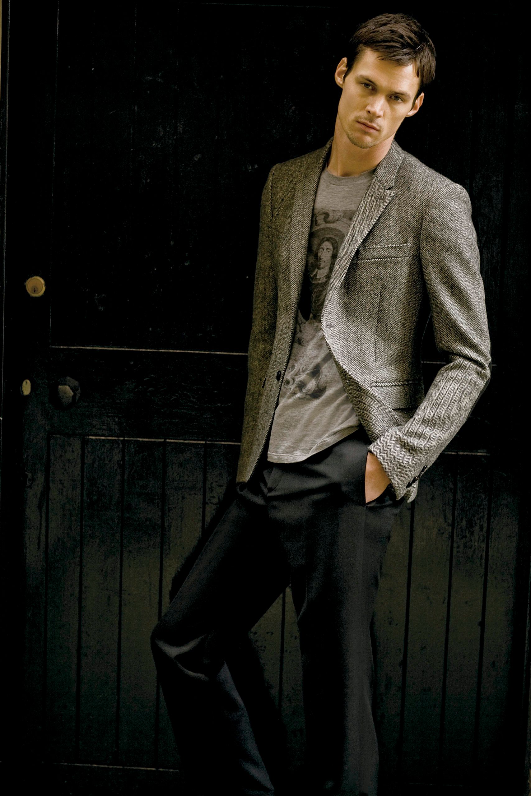 Man in grey tweed blazer and graphic tee with dark trousers leaning against a wooden door.