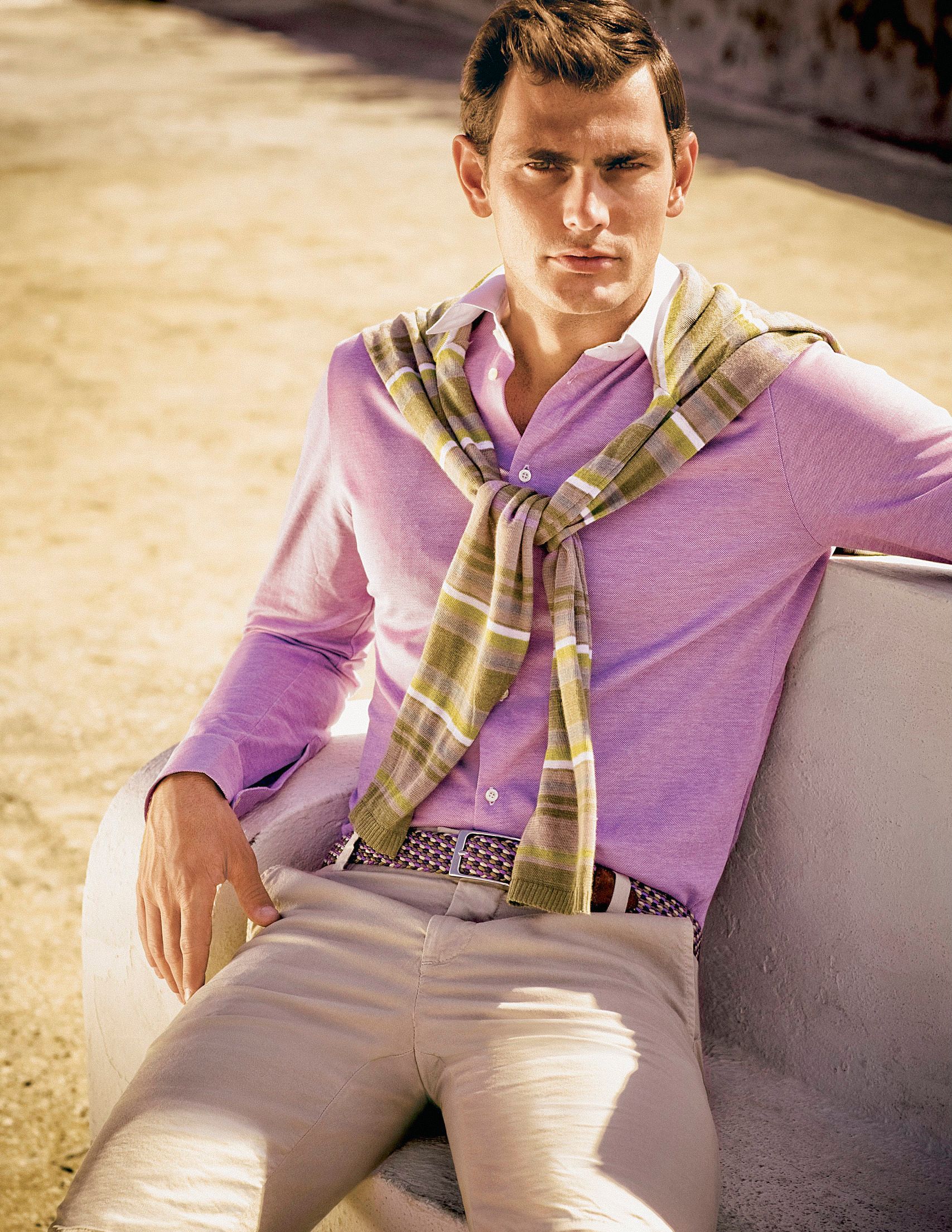 Man in pink sweater and plaid scarf seated outdoors in white pants, styled in a smart casual summer look.