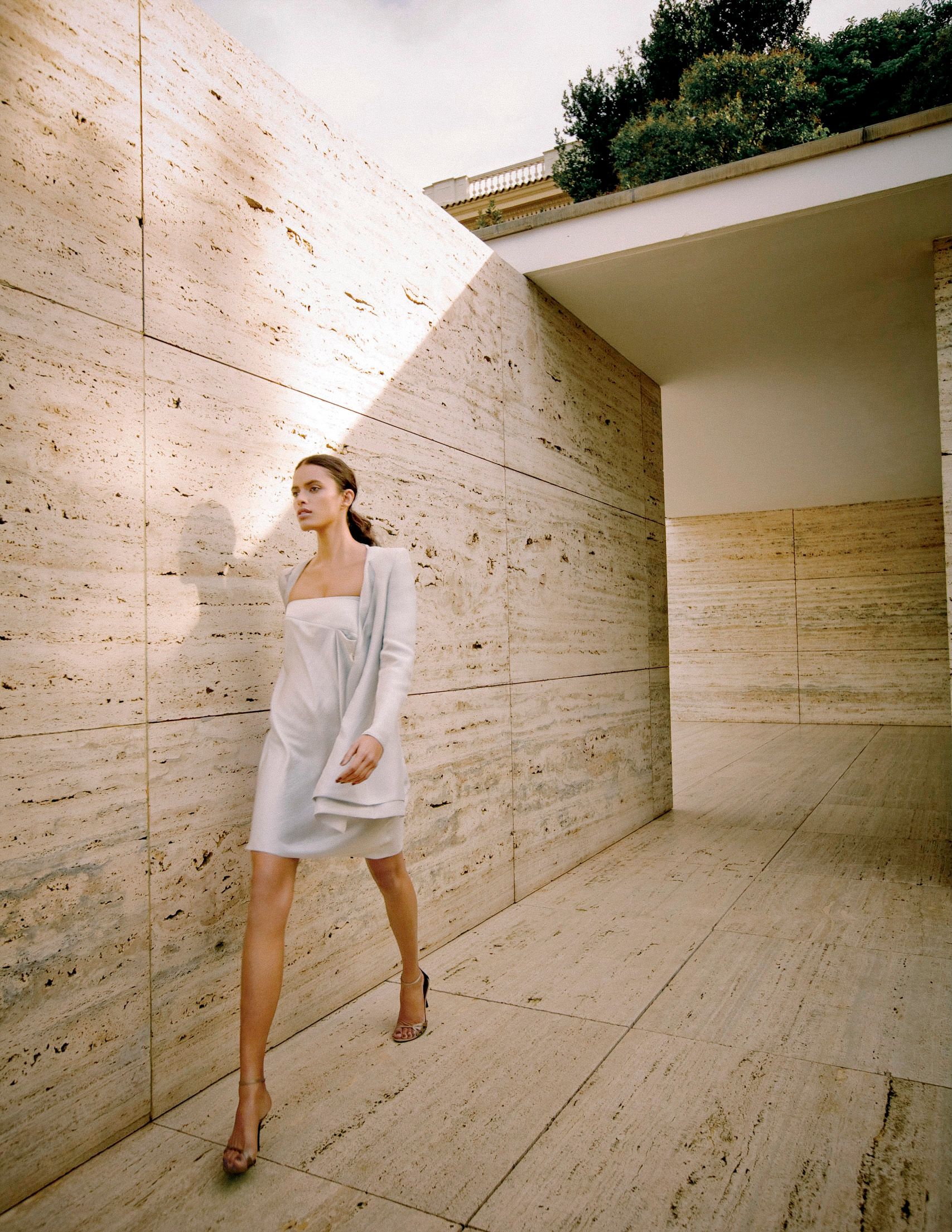 Woman in white mini dress and heels poses against travertine stone wall in modern architectural courtyard.