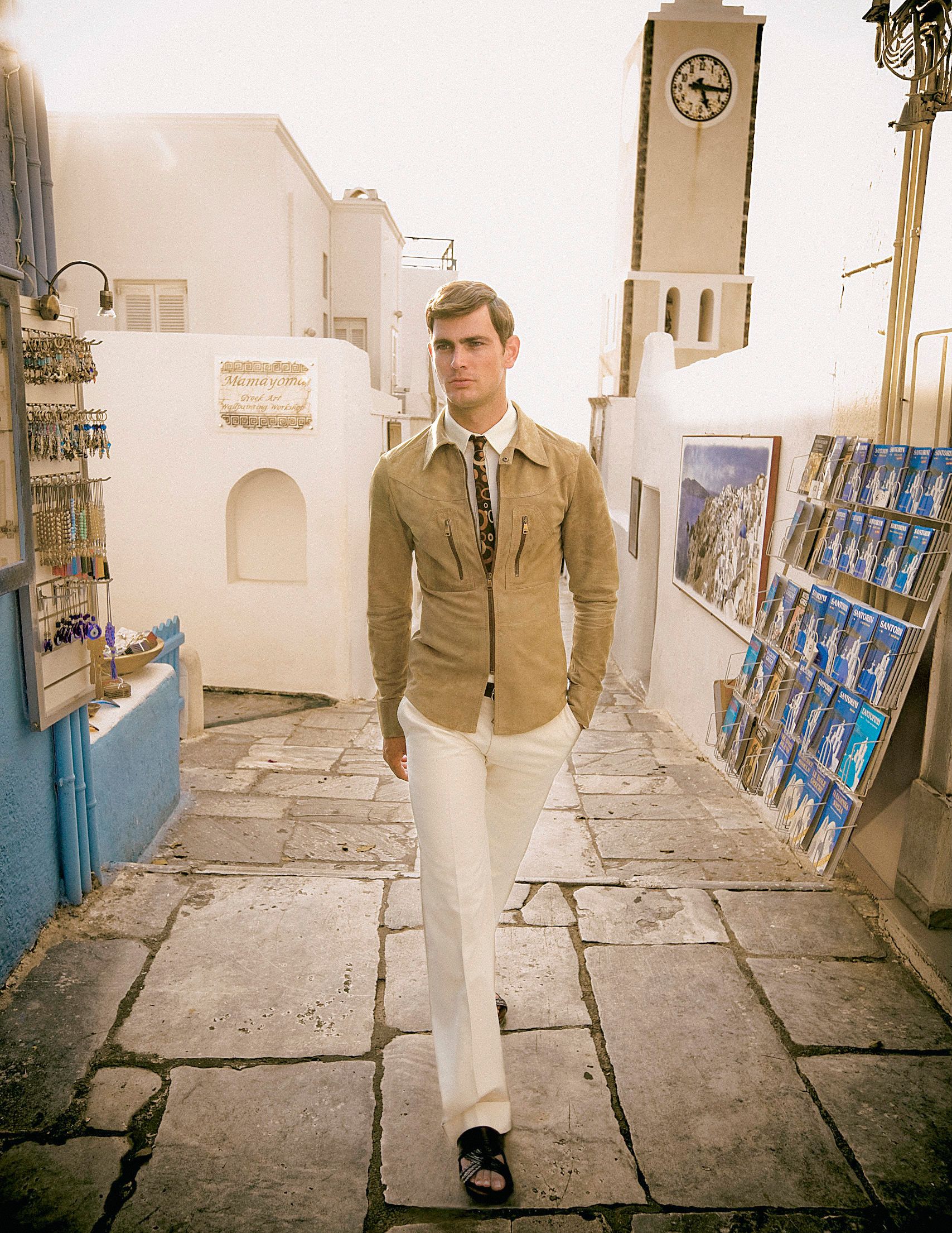 A stylish man walks through a Mediterranean alley in beige jacket and white trousers, with a clock tower and art displays nearby.
