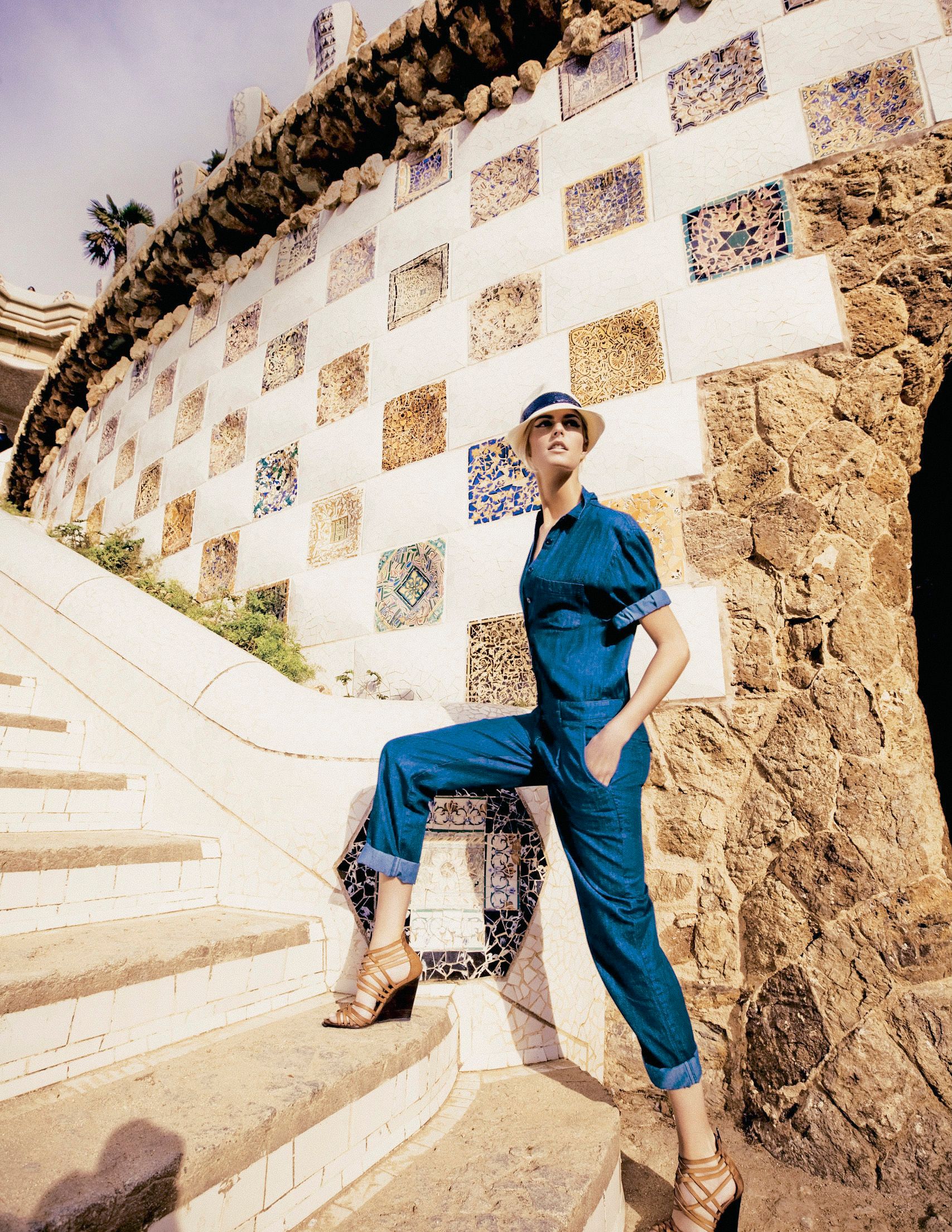 Woman in teal jumpsuit and hat posing on mosaic-tiled steps at Park Güell, Barcelona.