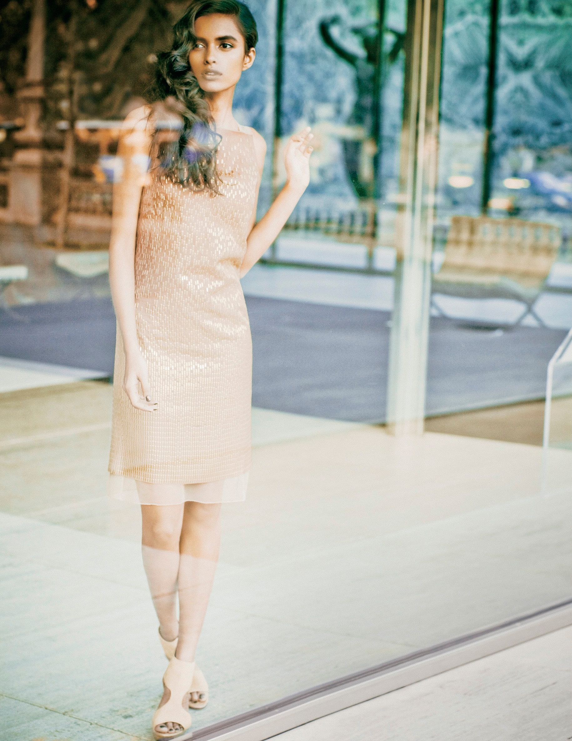 A model poses in a blush sequin layered dress with nude heels, standing near a glass window in natural light.