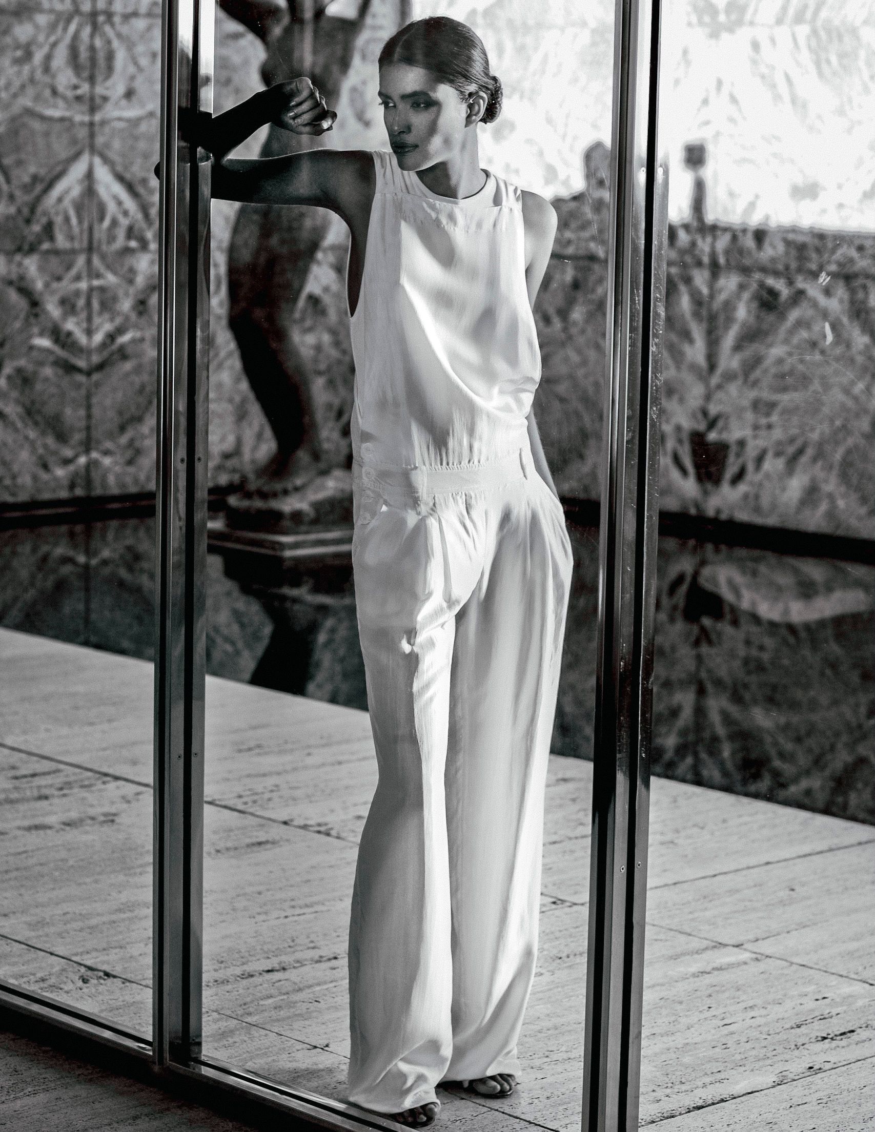 Black and white photo of a woman in elegant white wide-leg pants and sleeveless top posing by glass doors.