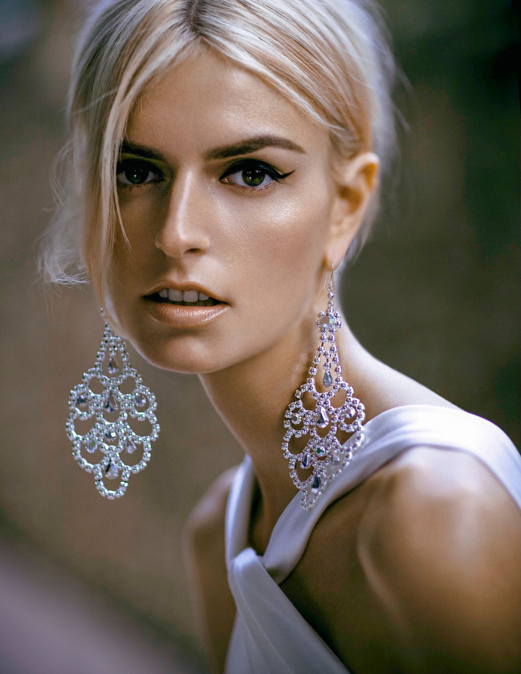 Blonde woman wearing large silver chandelier earrings and white dress, with elegant updo and dramatic eye makeup.
