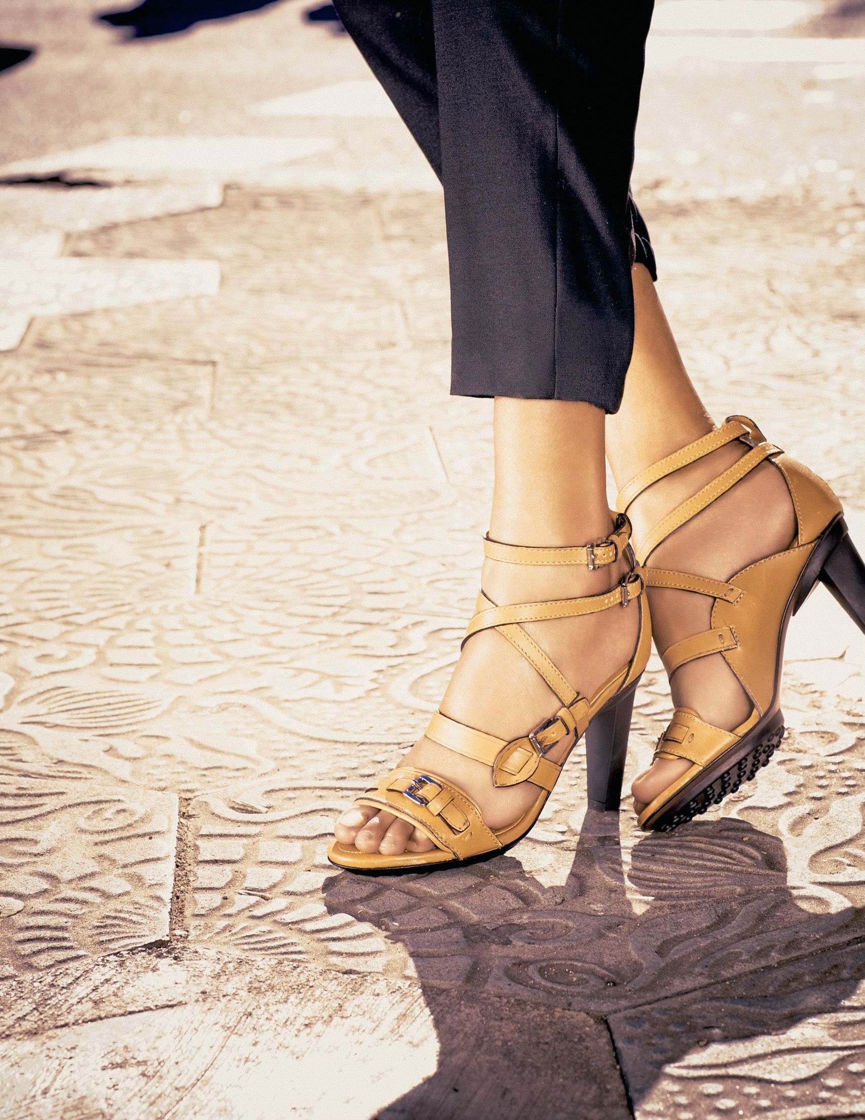 Woman wearing tan leather strappy high heel sandals with buckle details, paired with dark cropped trousers on a patterned surface.
