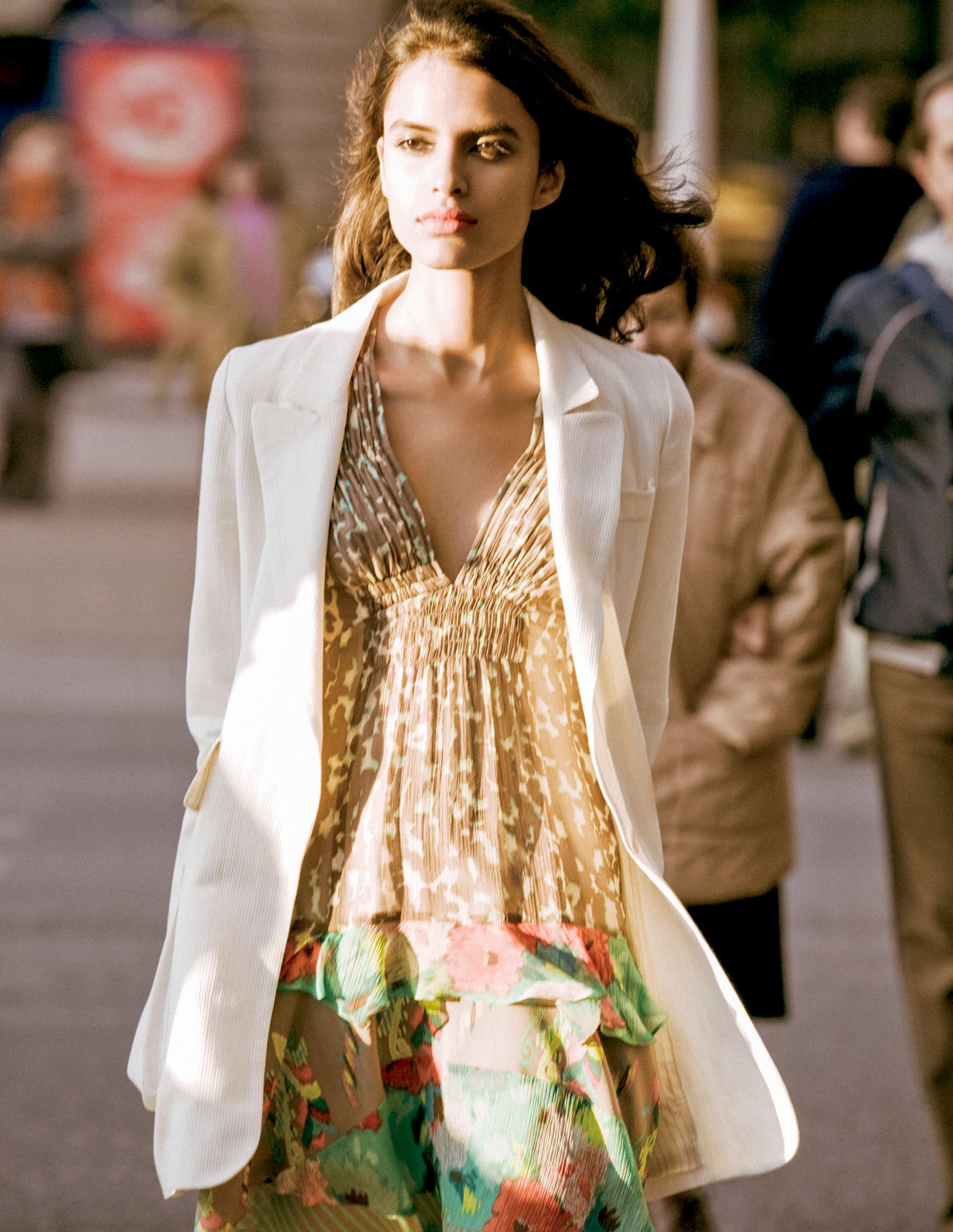 Woman in white blazer and floral printed dress walking outdoors in sunlight.
