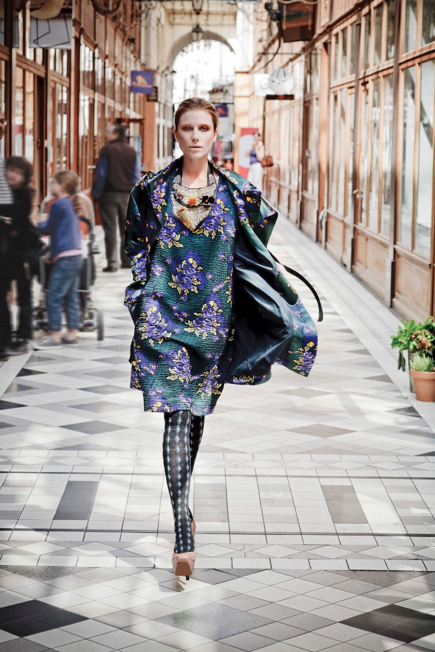 Model walks in floral brocade coat and patterned leggings through an elegant arcade gallery.