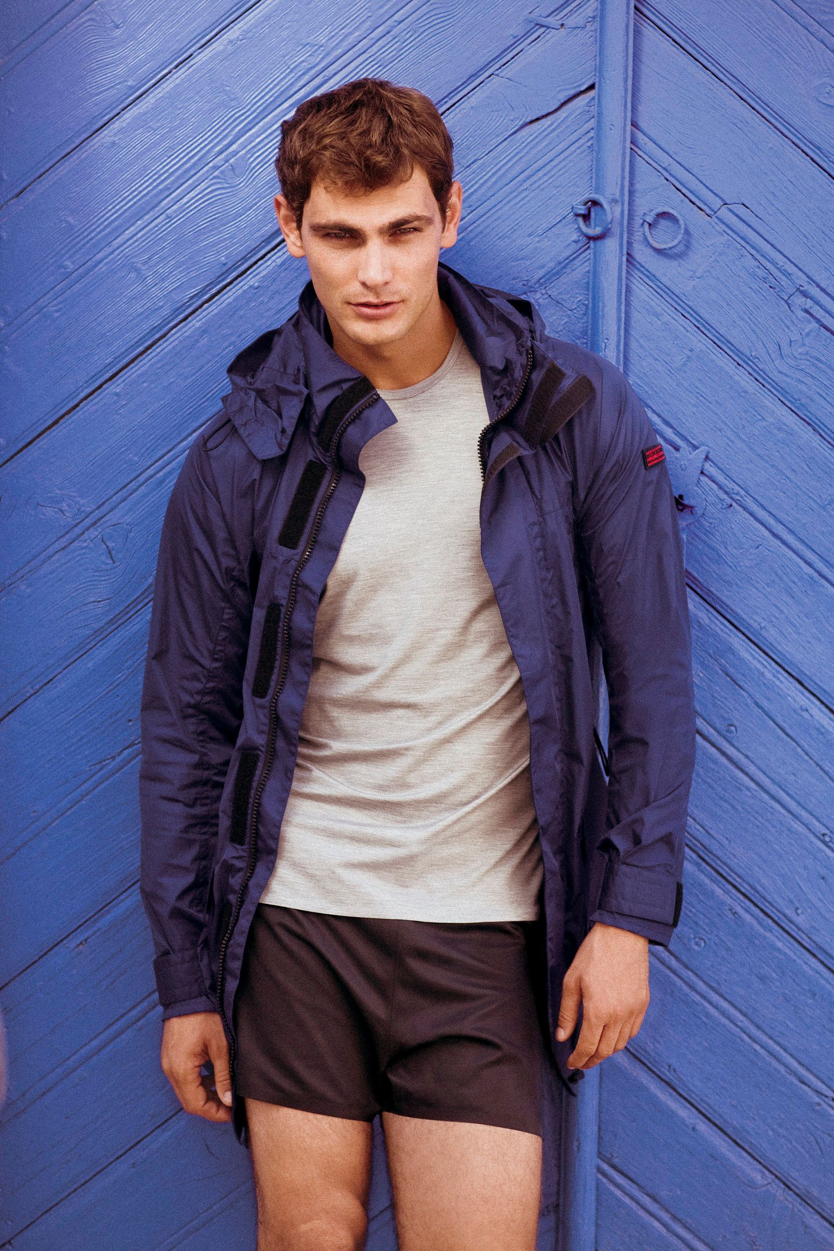 Young man in navy hooded jacket, grey t-shirt, and dark shorts leaning against a blue wooden wall.