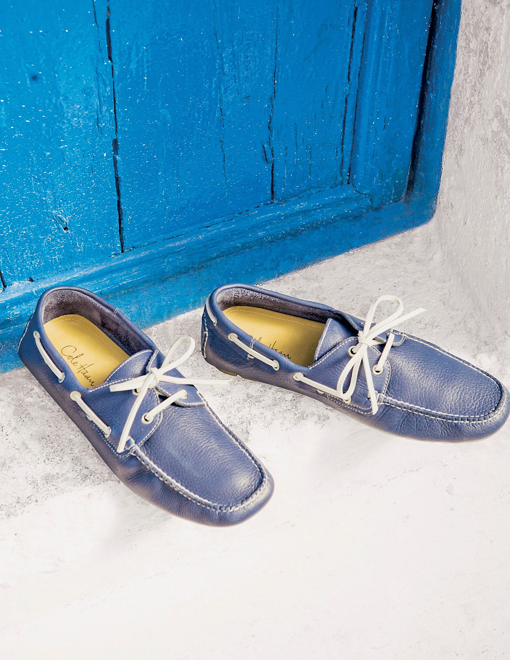 Blue leather boat shoes with white laces displayed against a vibrant blue wooden door on a white surface.